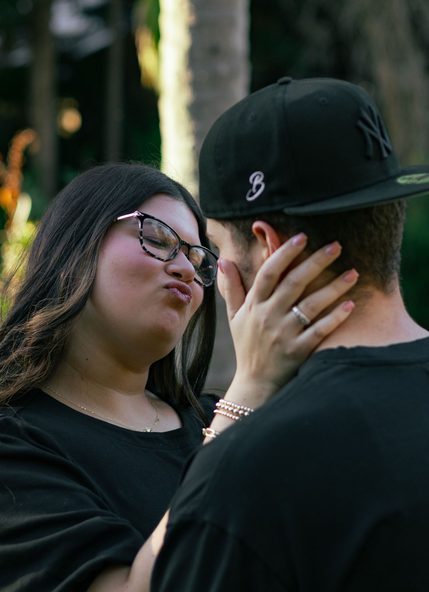 A woman with glasses playfully puckering lips toward her partner wearing a black NY cap in an outdoor setting.