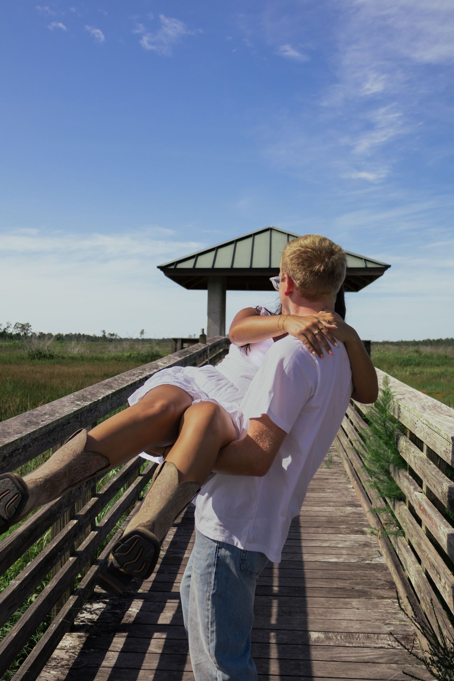 Man carrying woman in his arms on rustic wooden boardwalk with open sky and gazebo behind them.