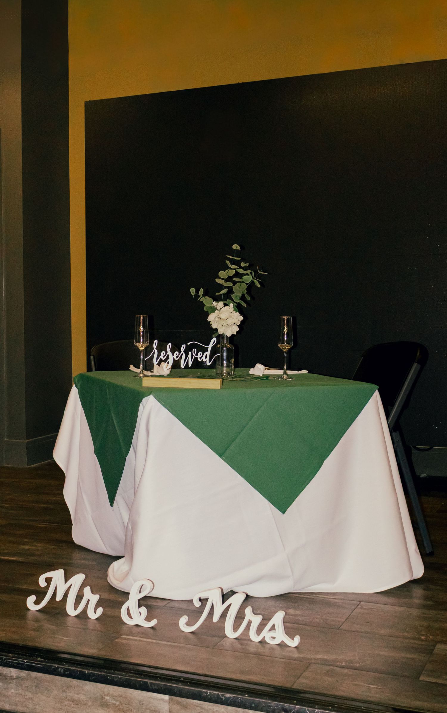Wedding sweetheart table with white and hunter green linens, floral centerpiece, and wooden Mr and Mrs letters on stage.