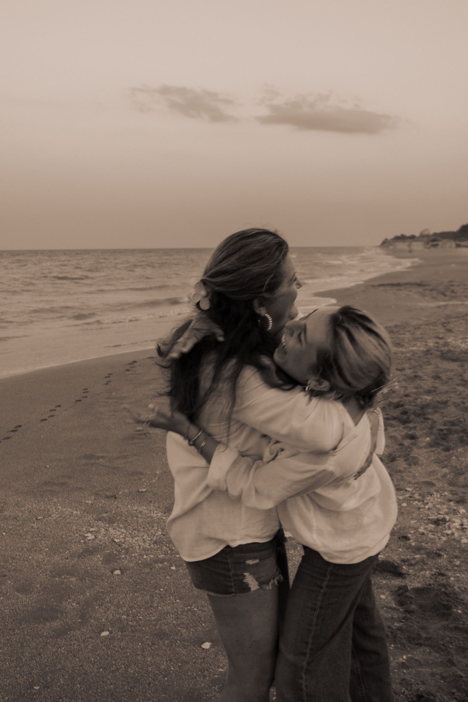 Two women sharing a warm embrace on a beach shoreline in a sepia-toned photo.