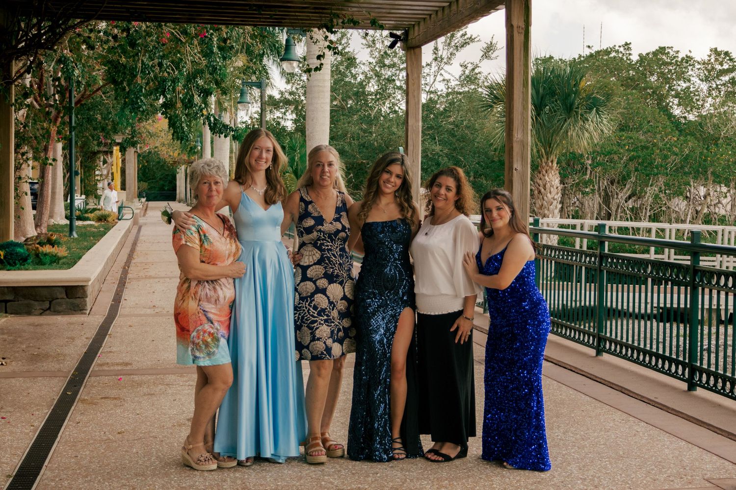 Group of six women in formal and casual attire pose together under a shaded outdoor pergola.