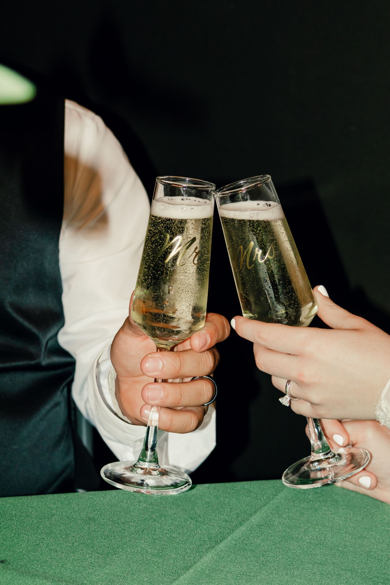 Close-up of bride and groom clinking Mr and Mrs champagne flutes over a green tablecloth at their wedding reception.