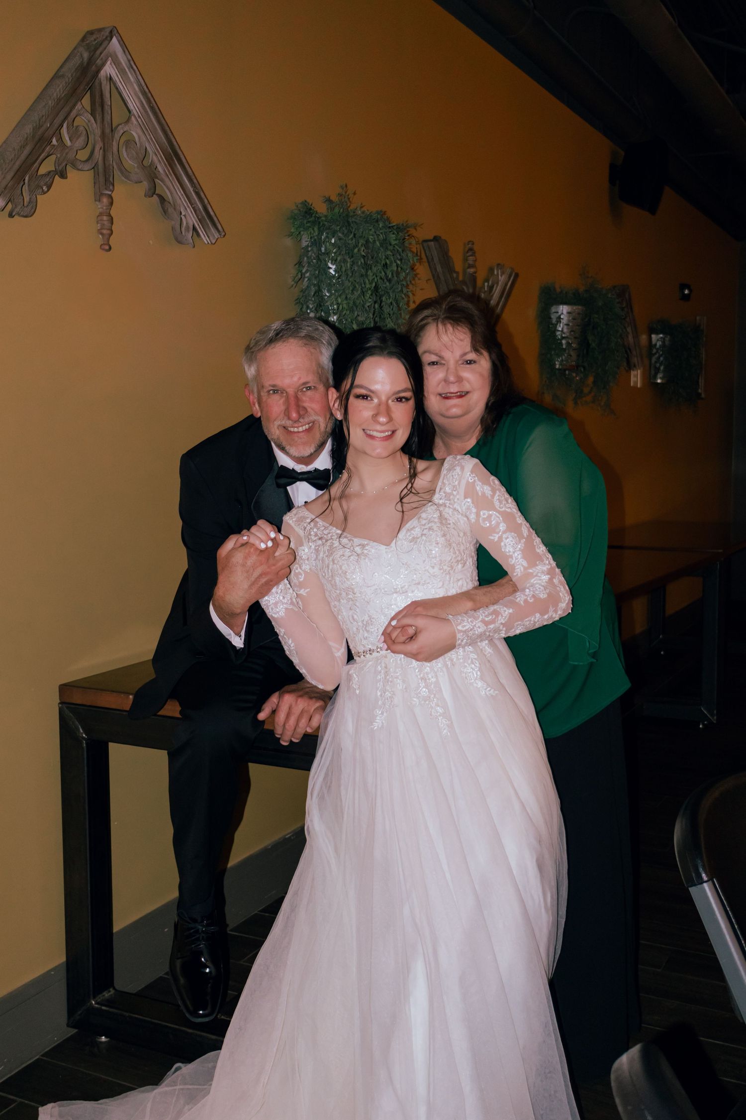 Bride in white lace gown poses with parents at elegant wedding reception venue.