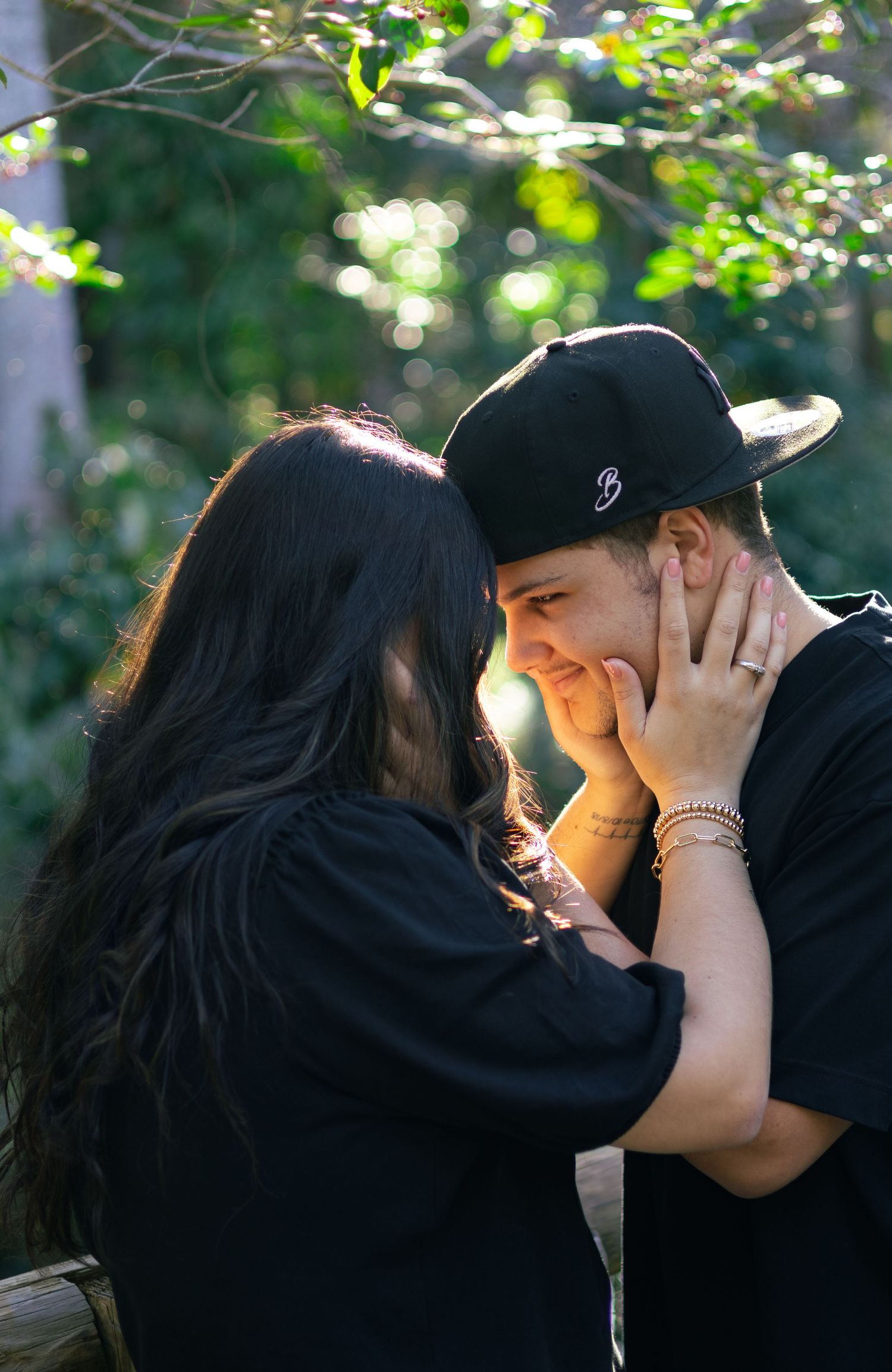 A couple in black sharing a tender moment outdoors, woman gently holding mans face as they lean close together.