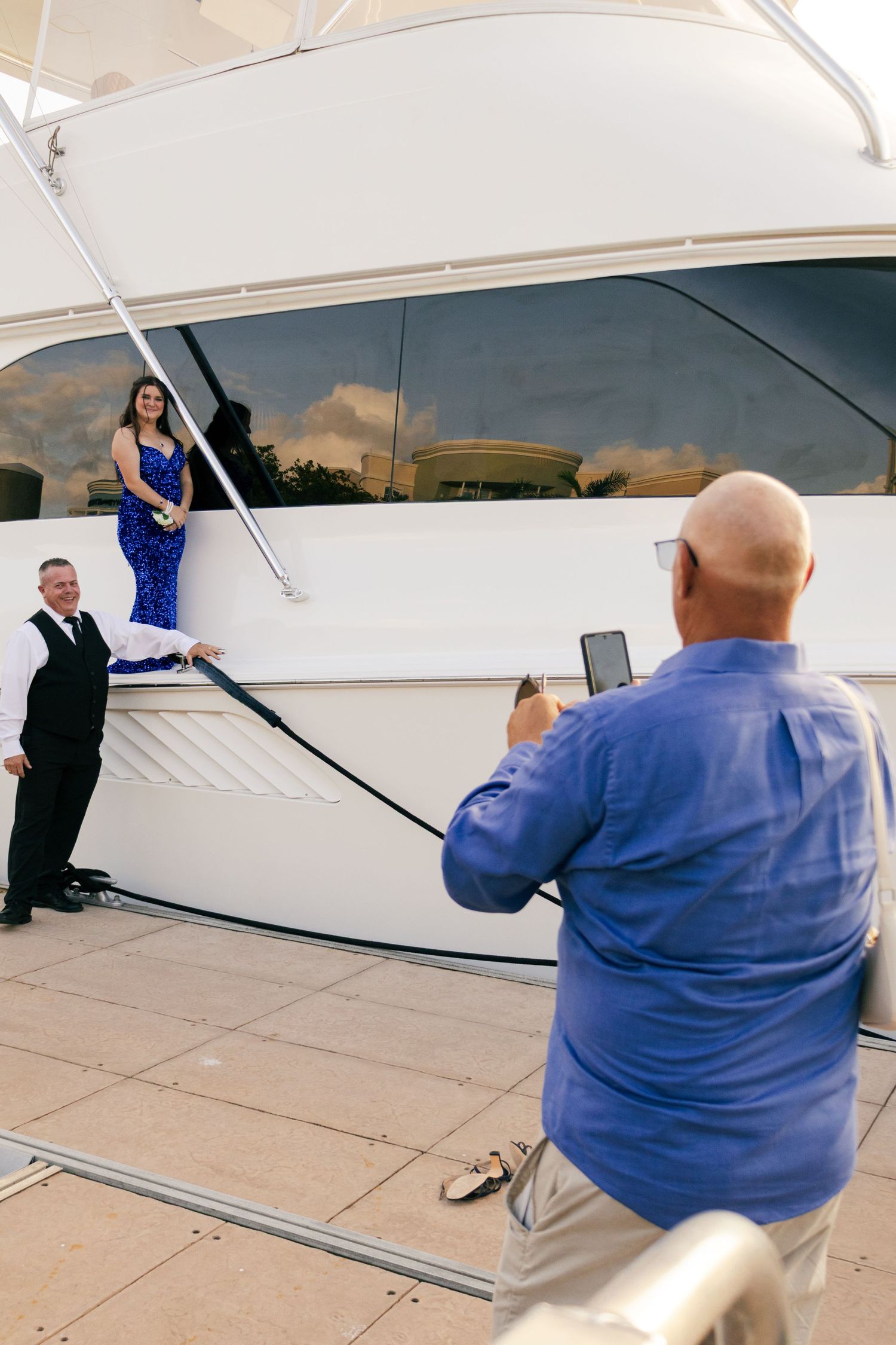 Man photographing couple in formal wear posing beside a large white luxury yacht at the dock.