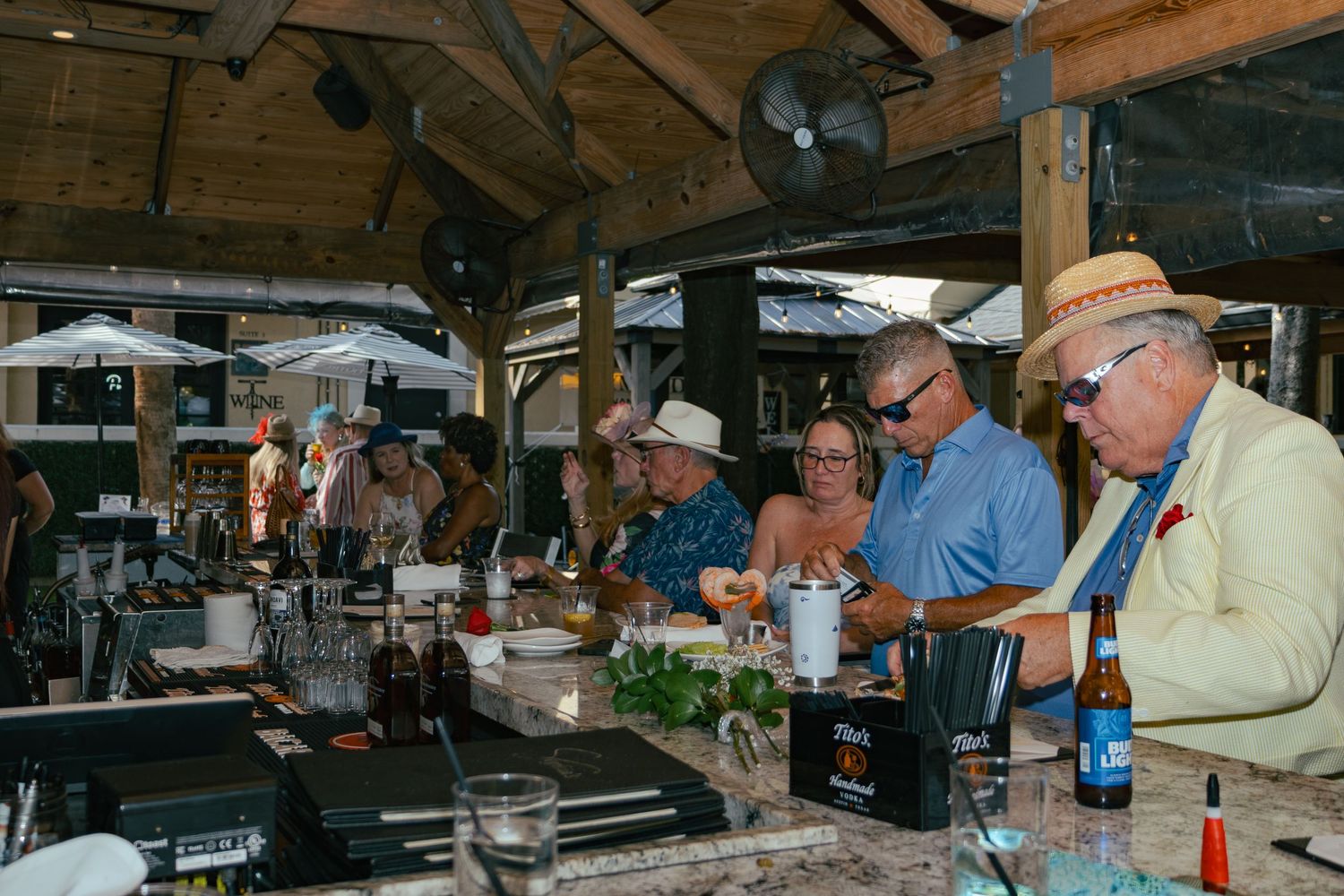 Guests in festive hats gather at outdoor bar during a Kentucky Derby-themed celebration.