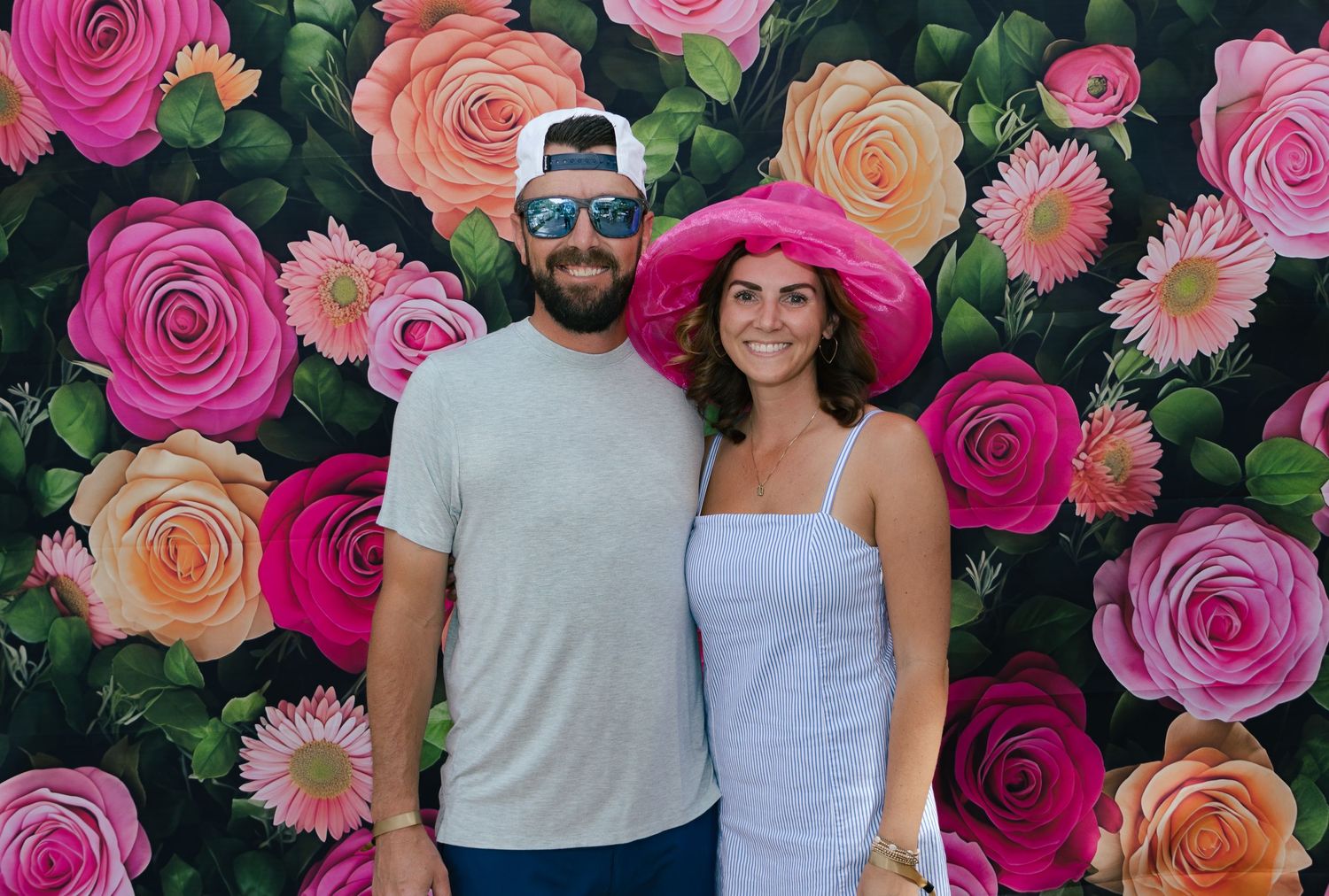 Couple posing in front of a colorful floral backdrop, woman wearing a large pink Kentucky Derby hat.