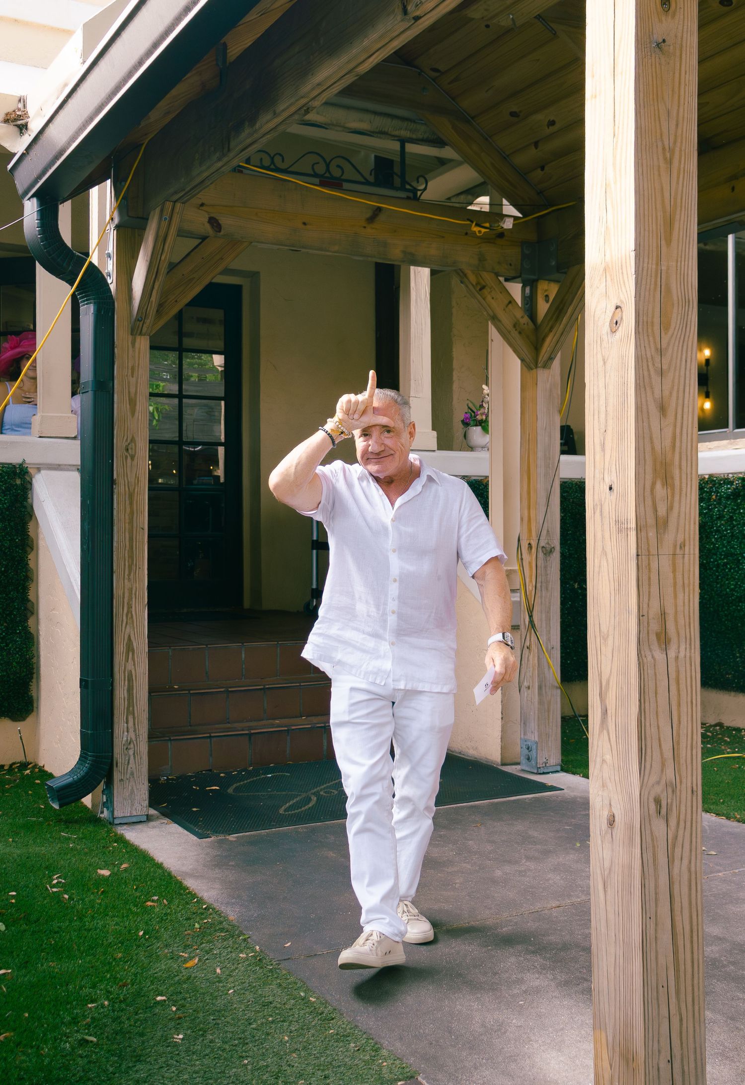 Man in white outfit walking under wooden pergola giving thumbs up gesture outdoors.