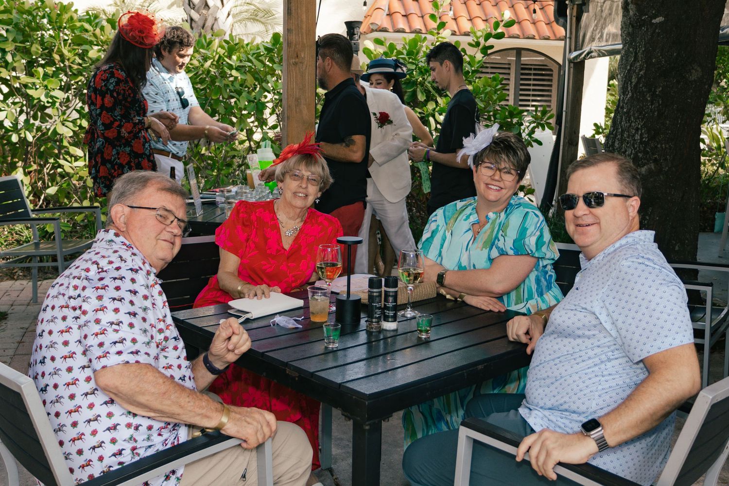 Four adults smile at an outdoor patio table during a sunny garden party event.
