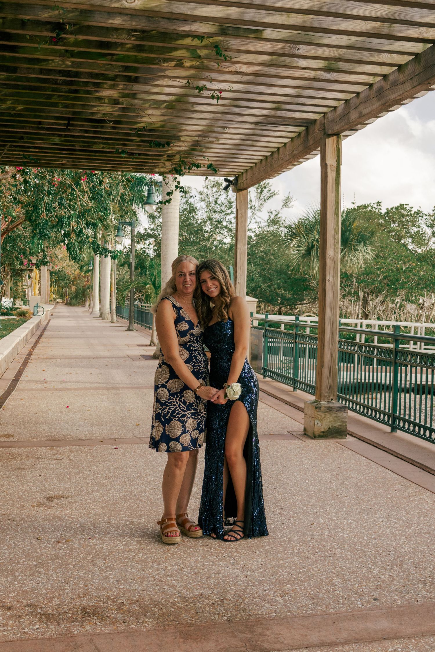 Two women posing together under a rustic wooden pergola, one wearing a floral dress and one in a sequin gown.