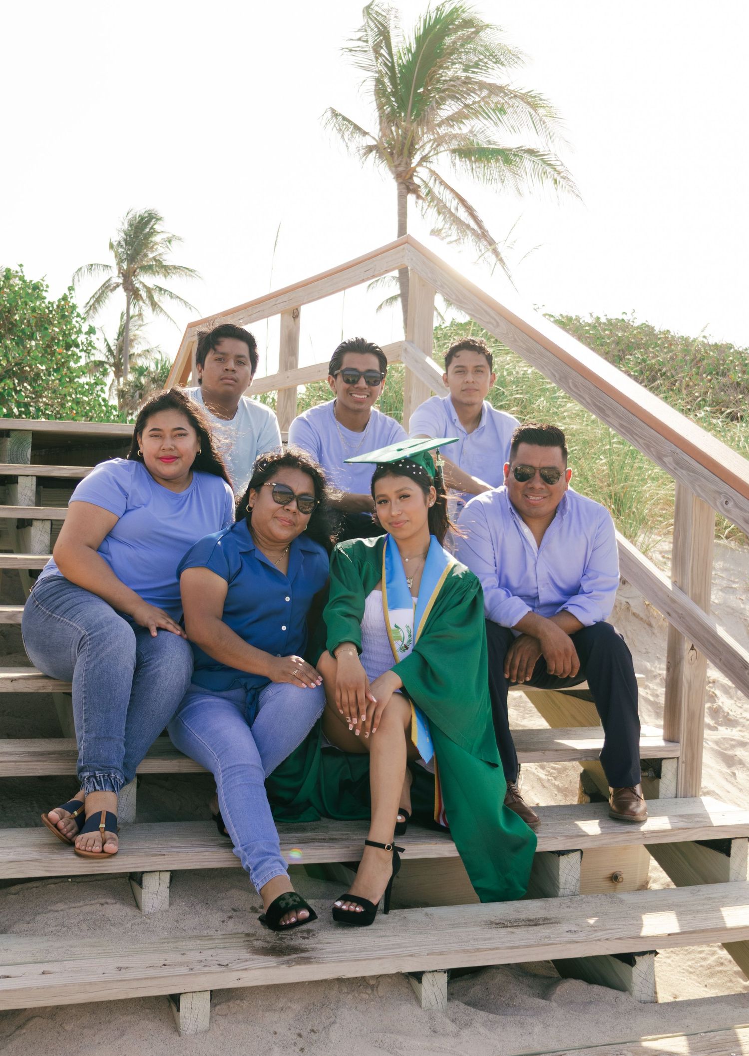 Family celebrates graduation on beach steps, graduate in green cap and gown surrounded by proud family members with palm trees.