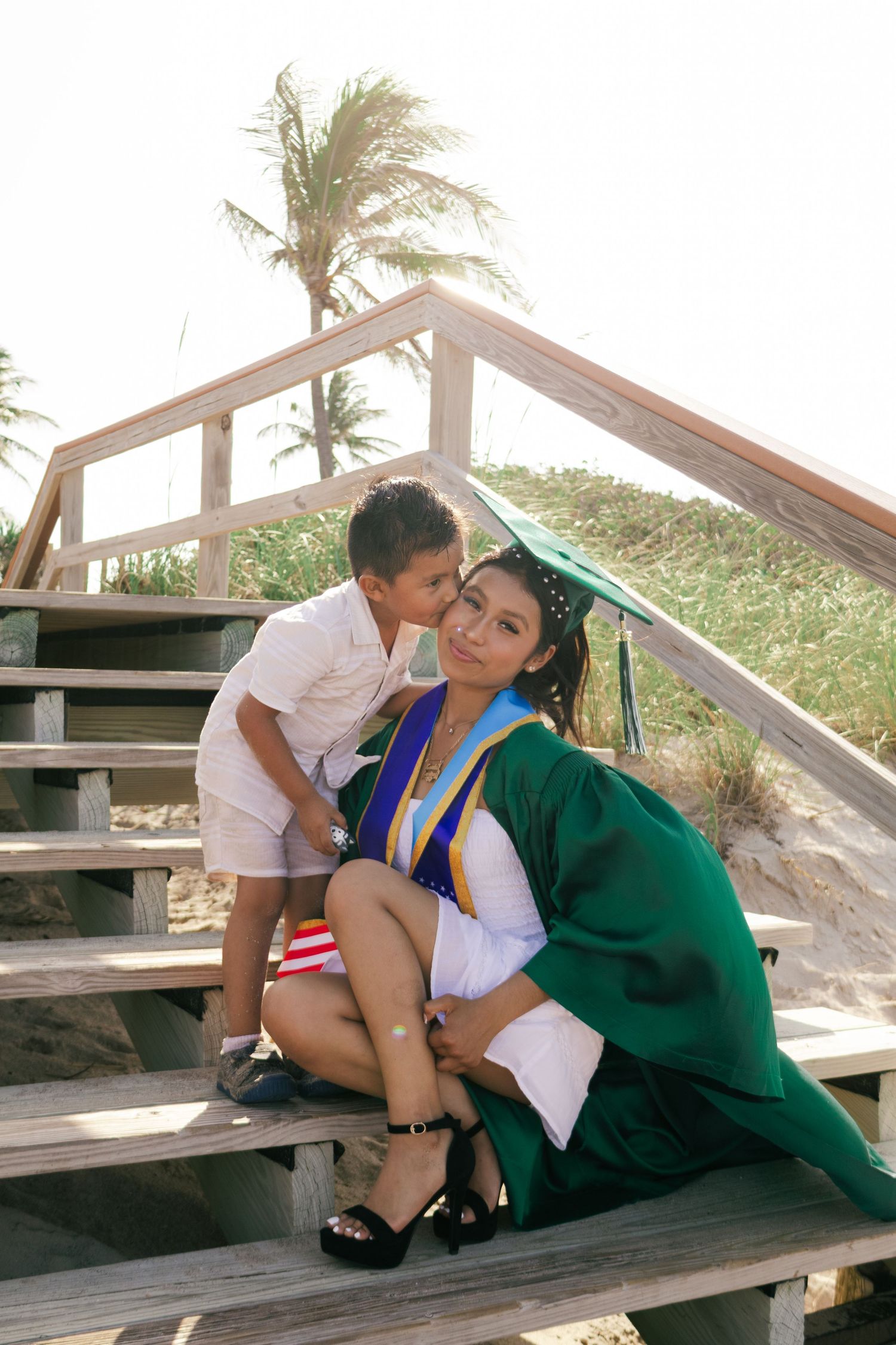Young graduate in green cap and gown receiving a sweet kiss on the cheek from a small boy on beach stairs.