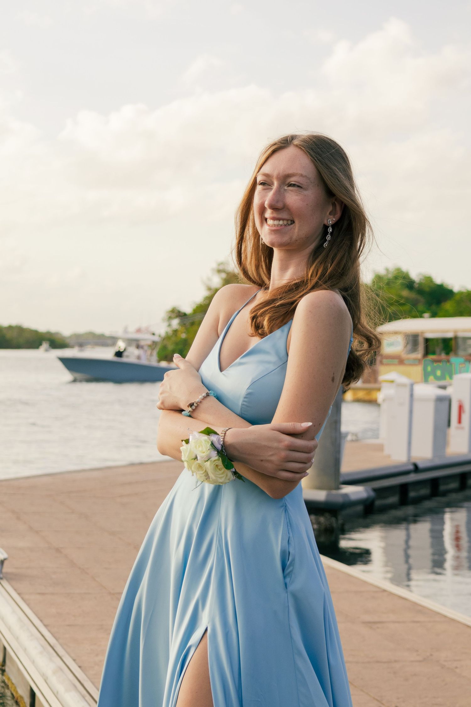 A teenage girl in a light blue prom dress laughs while standing on a waterfront dock with boats in background.