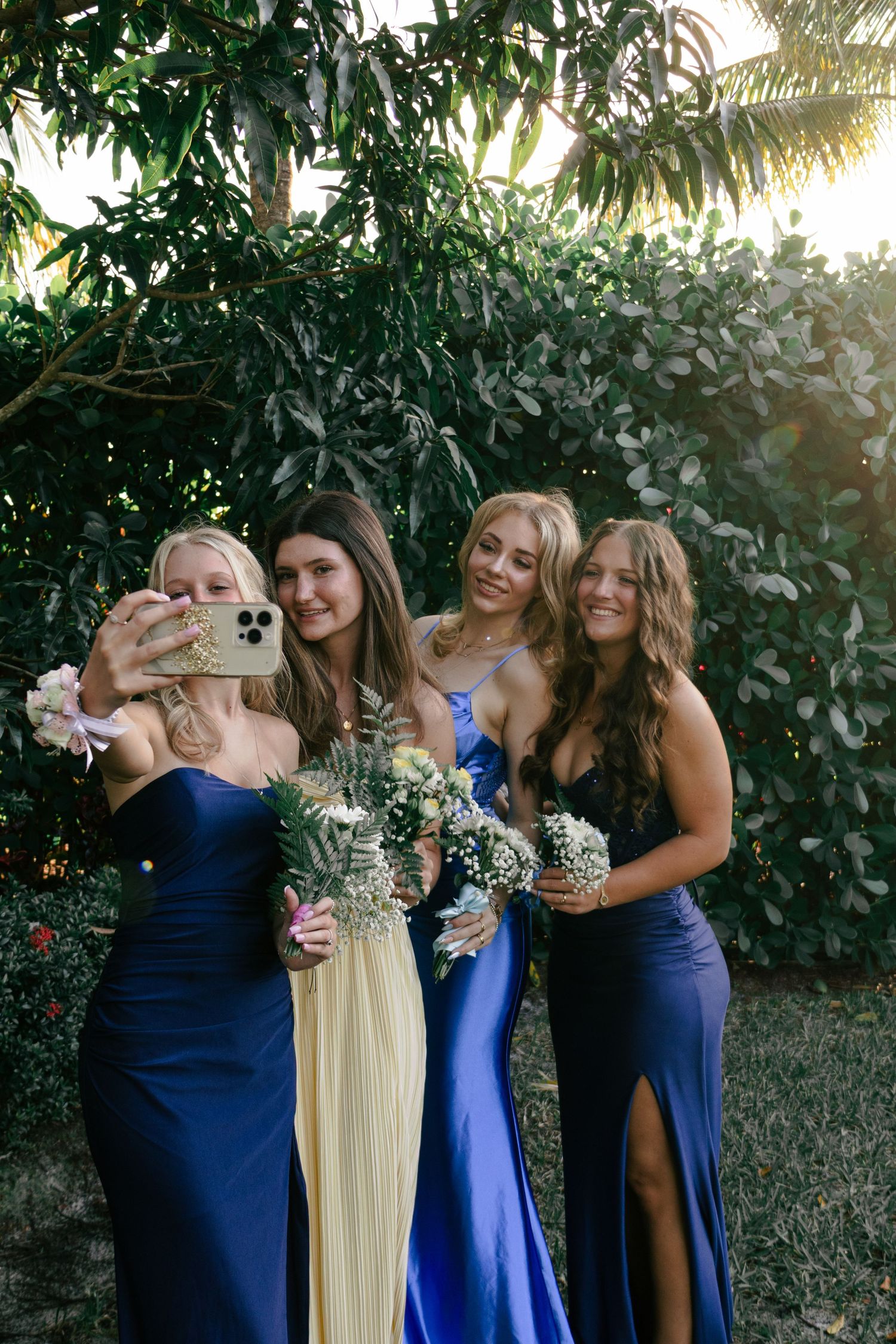 Four young women in formal gowns take a selfie outdoors while holding floral bouquets at prom.