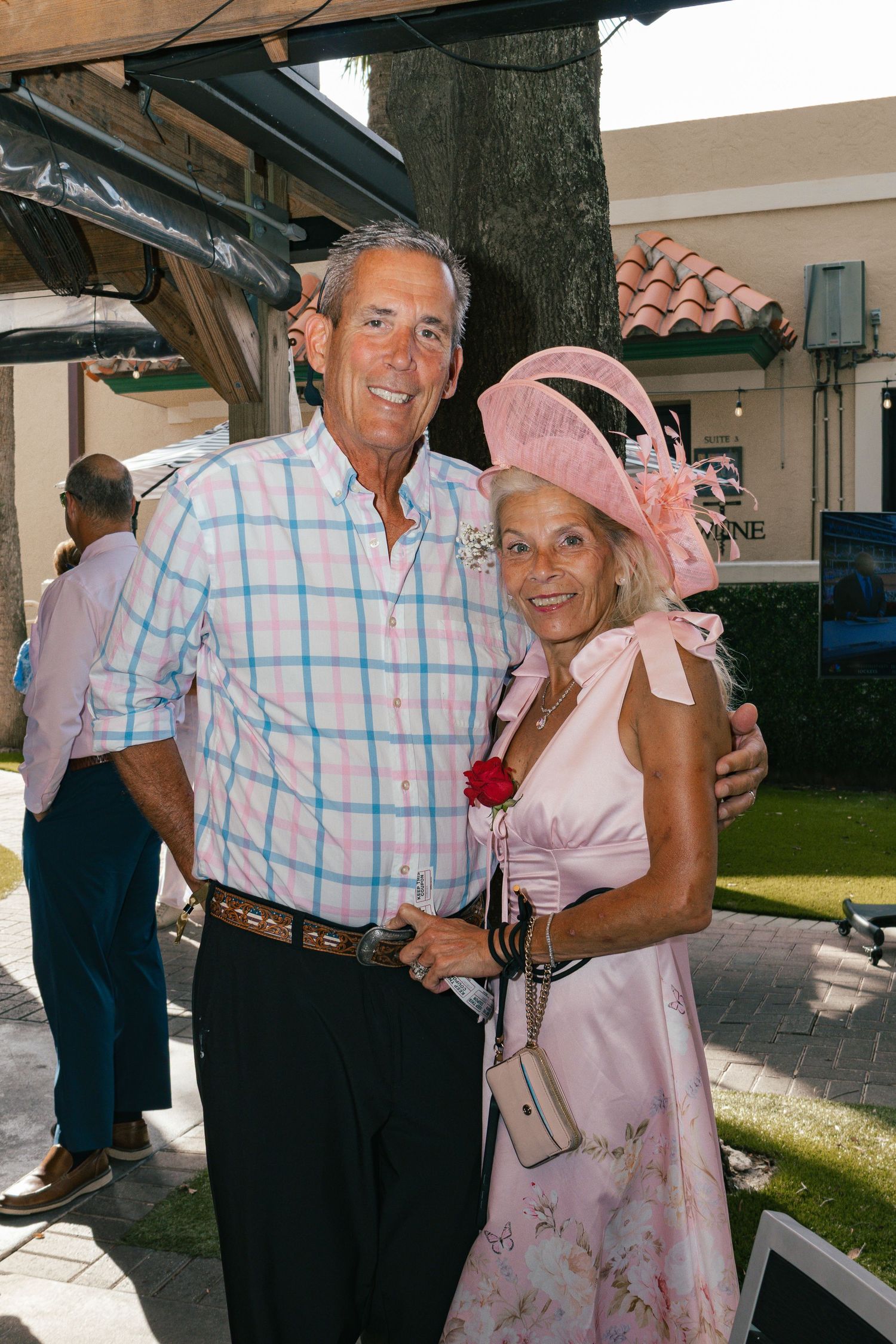 Smiling couple poses together at outdoor event, woman wearing pink dress and large pink fascinator hat.