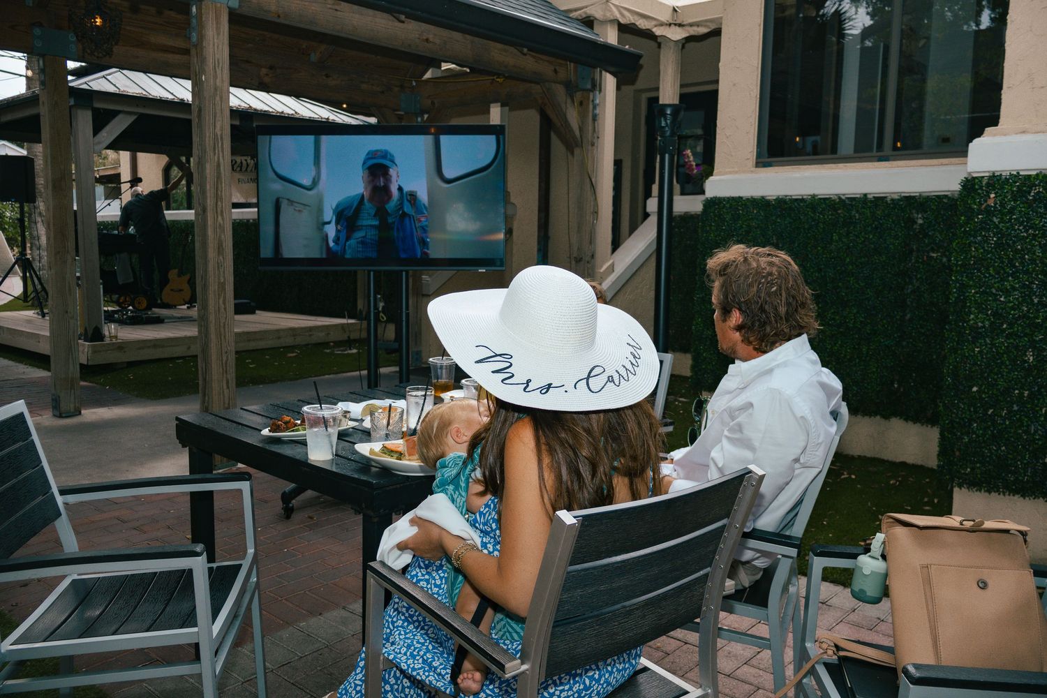 Couple in Derby attire watches race on outdoor screen, woman wearing white hat inscribed Mare-Gaux.