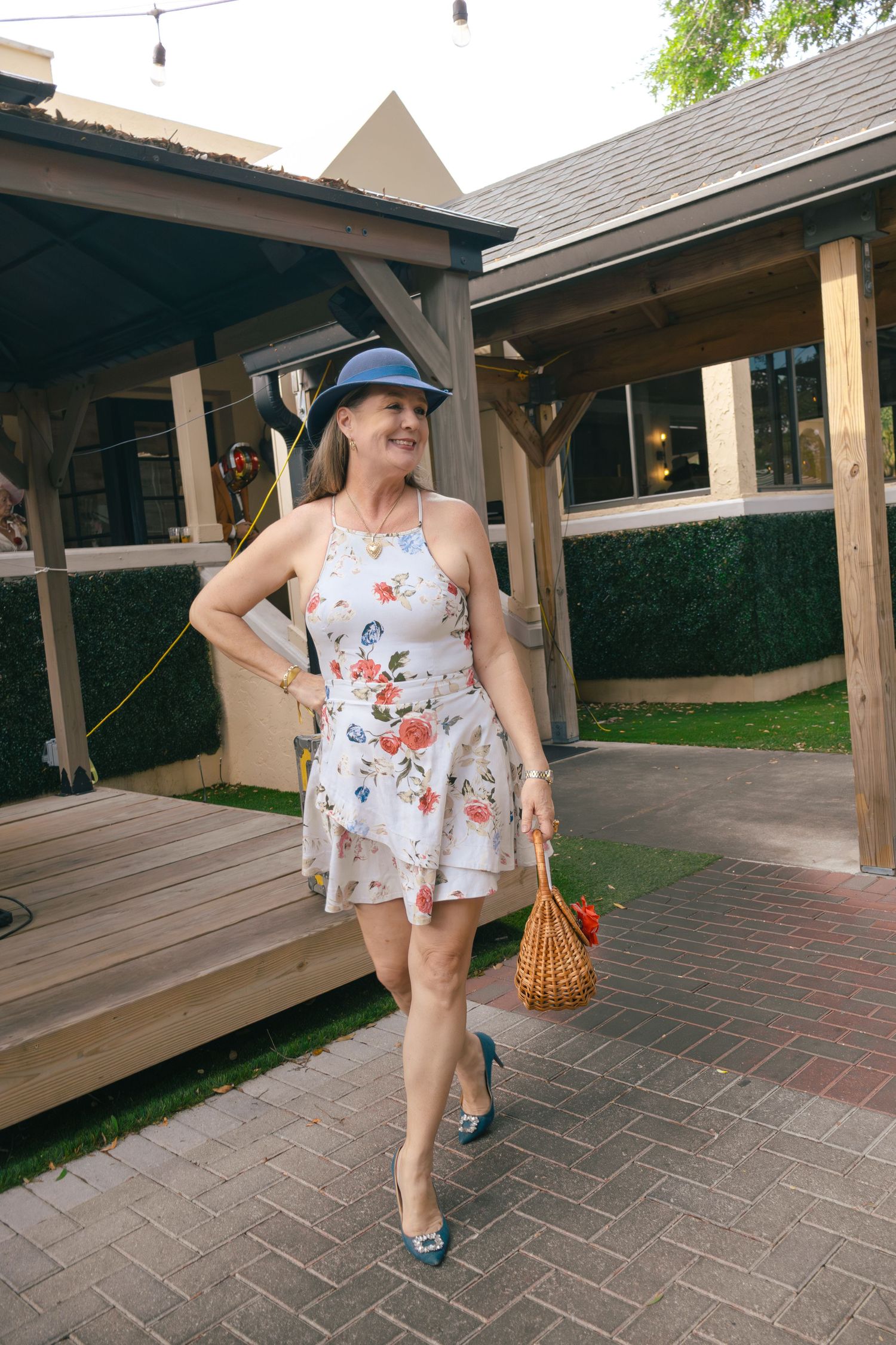 Woman in floral dress and blue hat walks through outdoor venue carrying wicker bag.