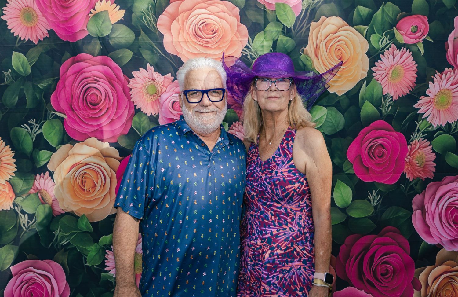 Couple in festive attire with purple hat posing before a vibrant painted floral rose wall backdrop.