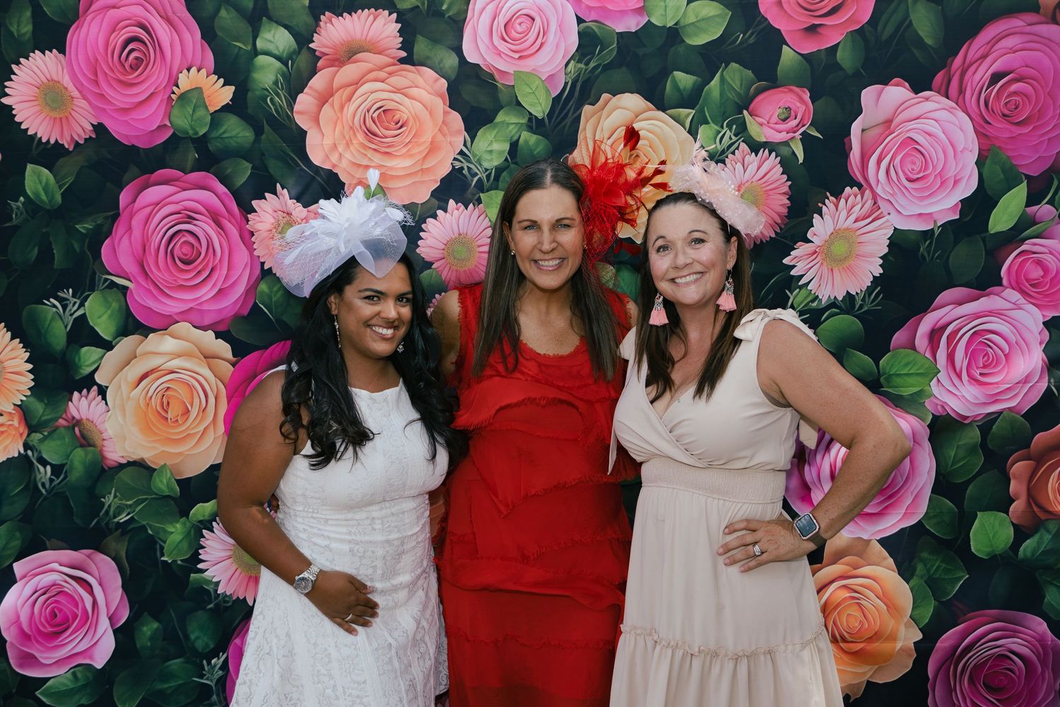 Three women in elegant dresses and fascinators pose in front of a colorful floral backdrop at a Kentucky Derby party.