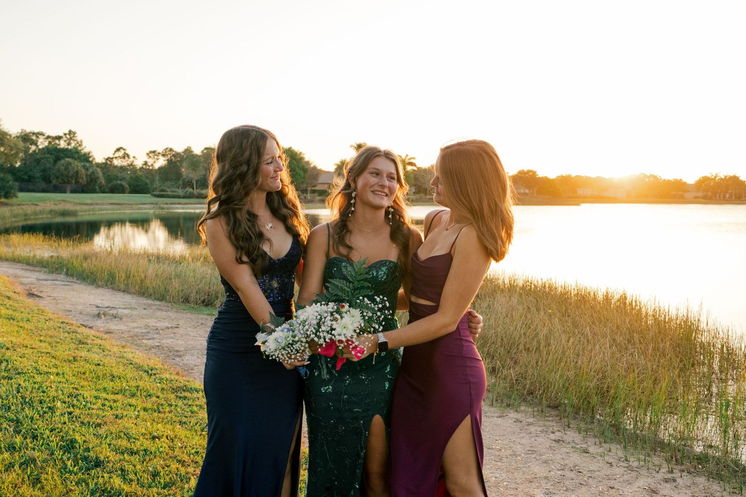 Three smiling young women in formal prom dresses pose together near a scenic lake at sunset.