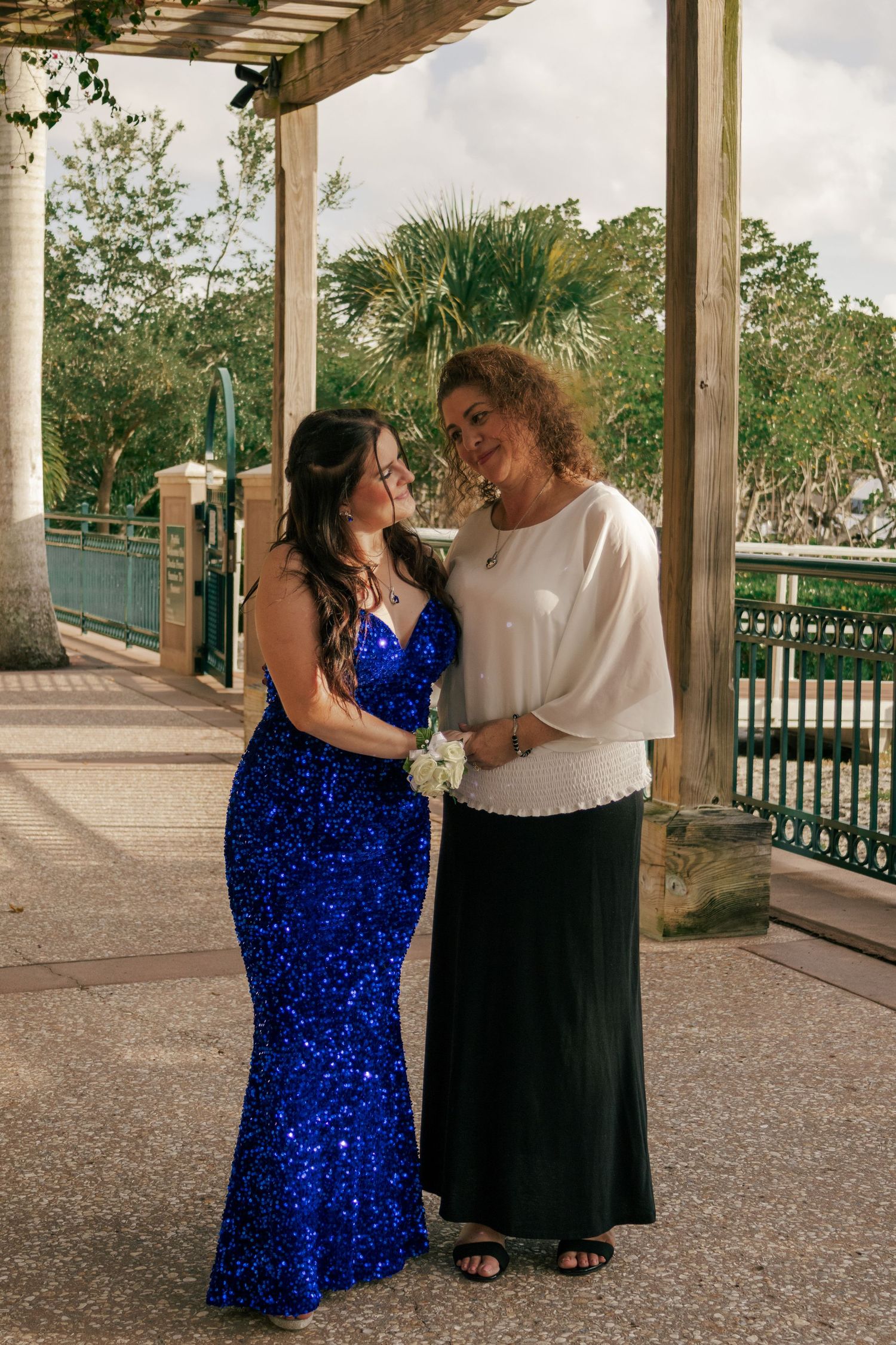 Young woman in a royal blue sequin gown standing with an older woman under a park pergola.