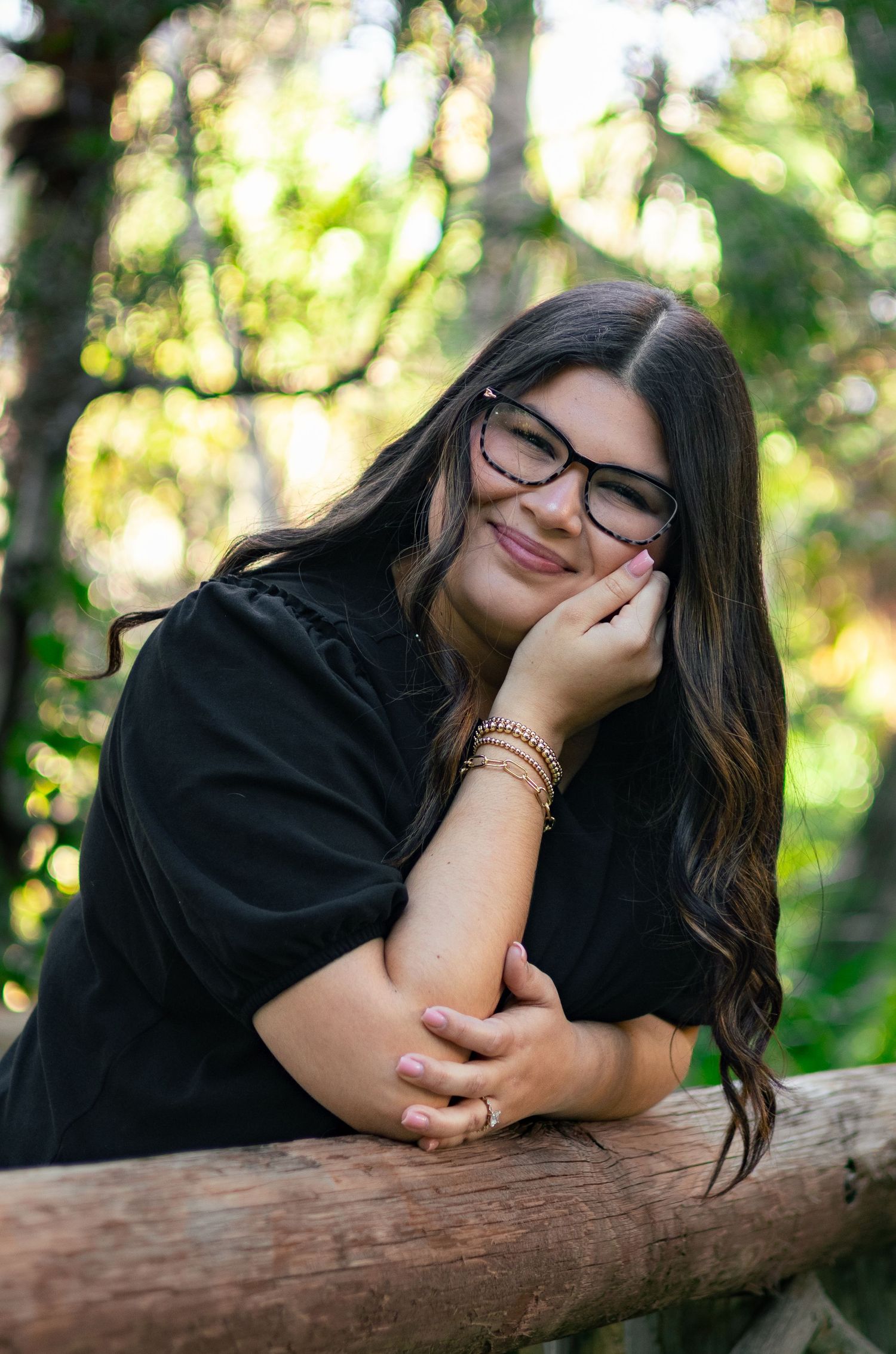 Smiling young woman with glasses resting on wooden log surrounded by green foliage outdoors.