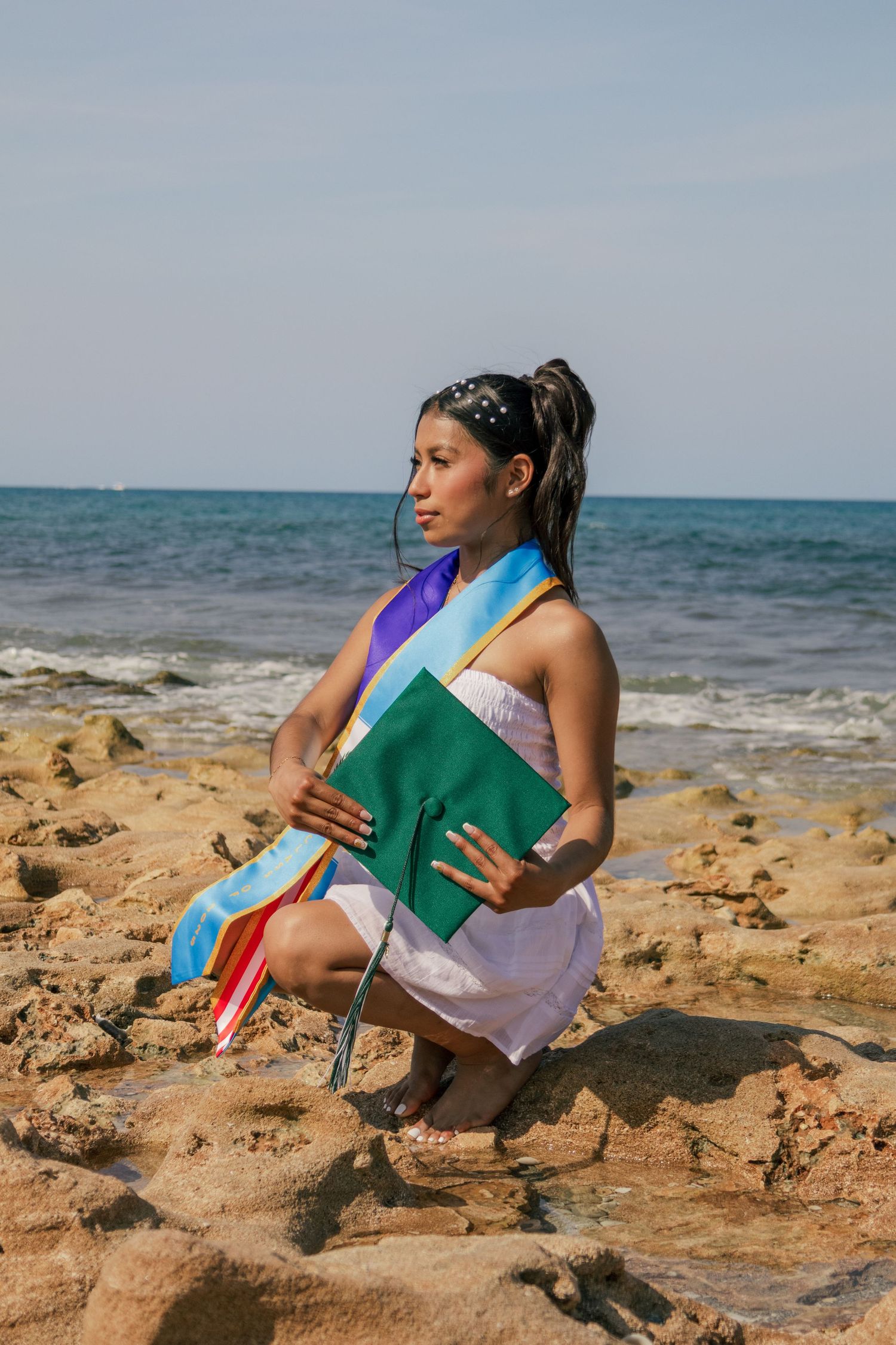 Young woman graduate sitting on rocky beach holding green mortarboard with colorful sashes.