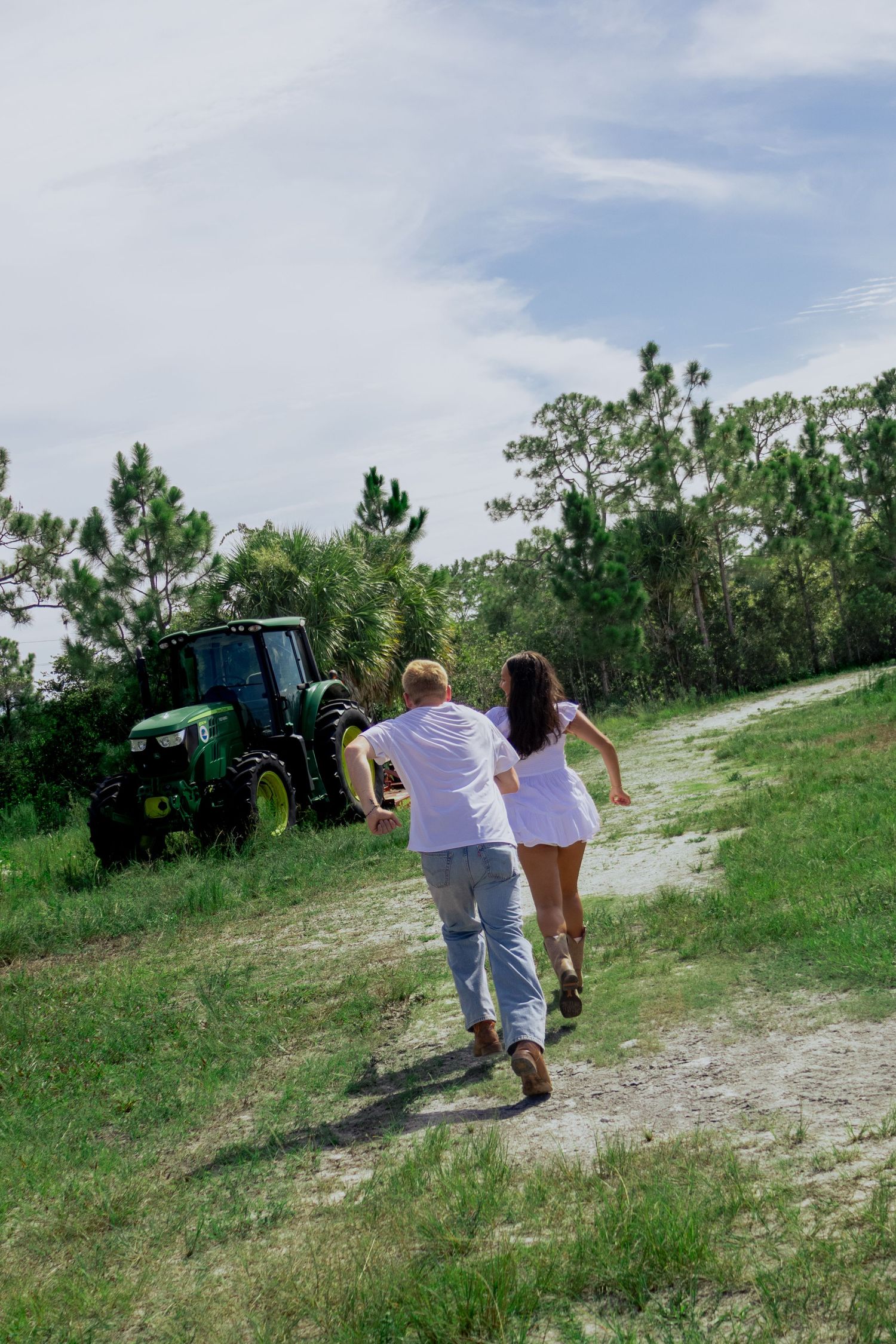 Couple running hand-in-hand down a rural dirt path toward a green John Deere tractor.