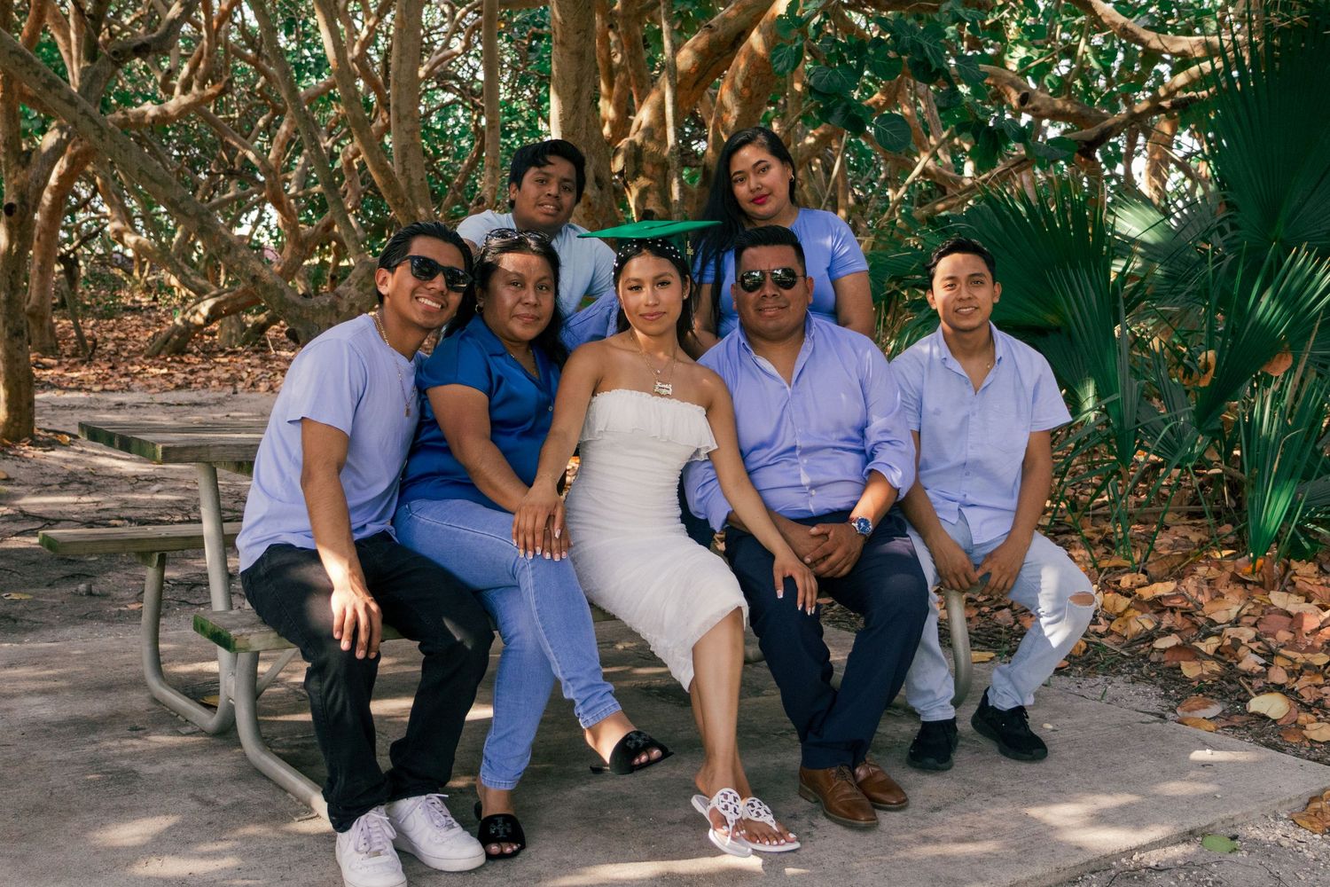 Group of seven people posing together on a bench surrounded by lush green trees in a park.