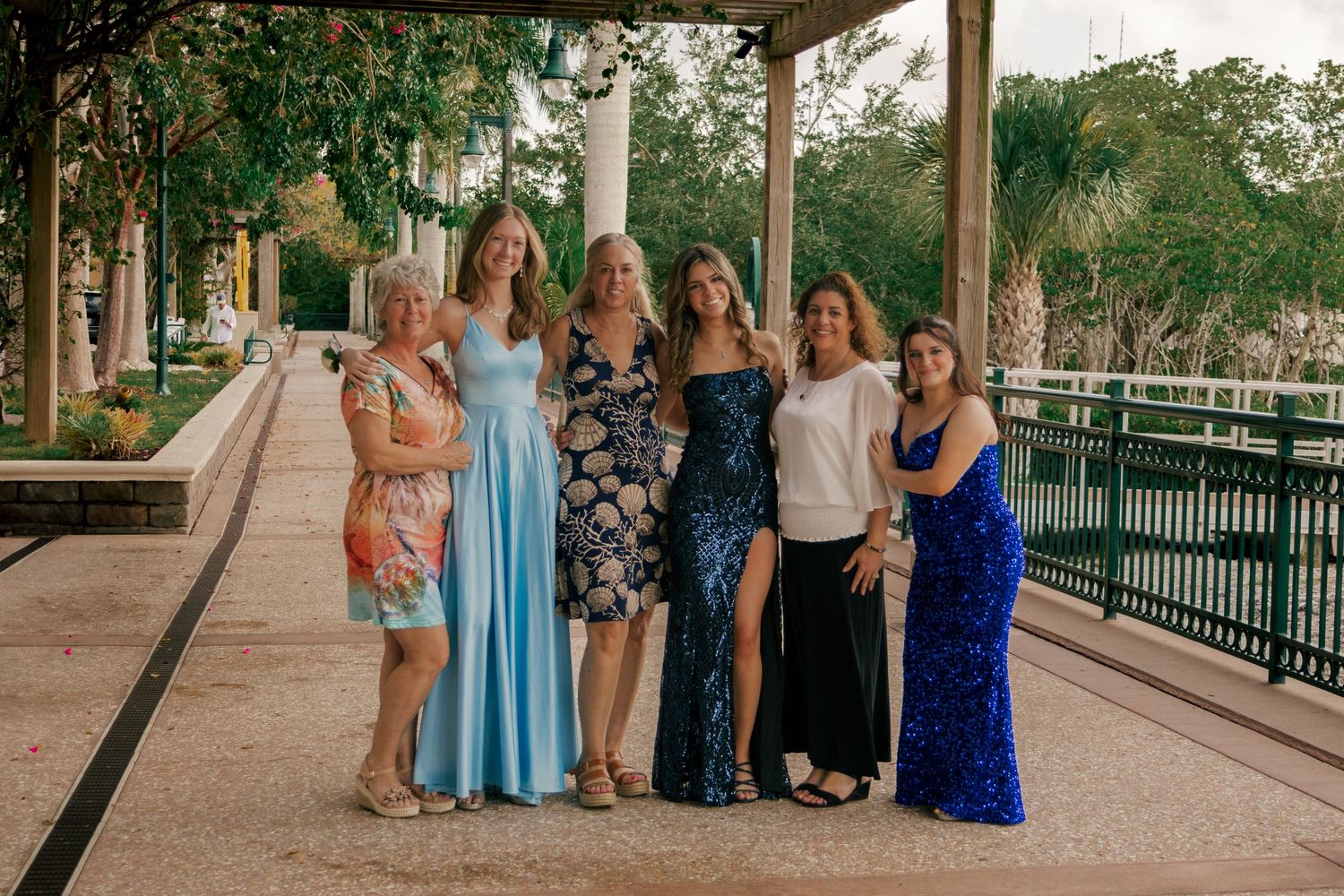 Six women in formal dresses pose together under a covered walkway at an outdoor venue.