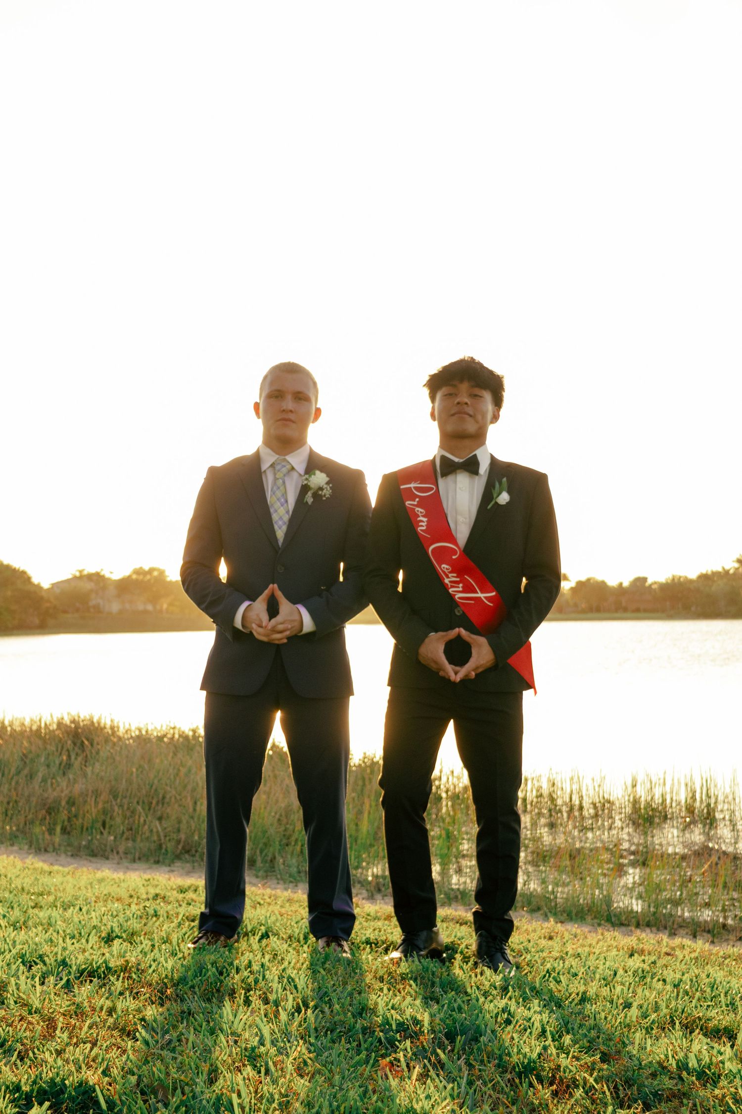 Two young men in formal suits pose outdoors at golden hour, one wearing a red Prom Court sash.