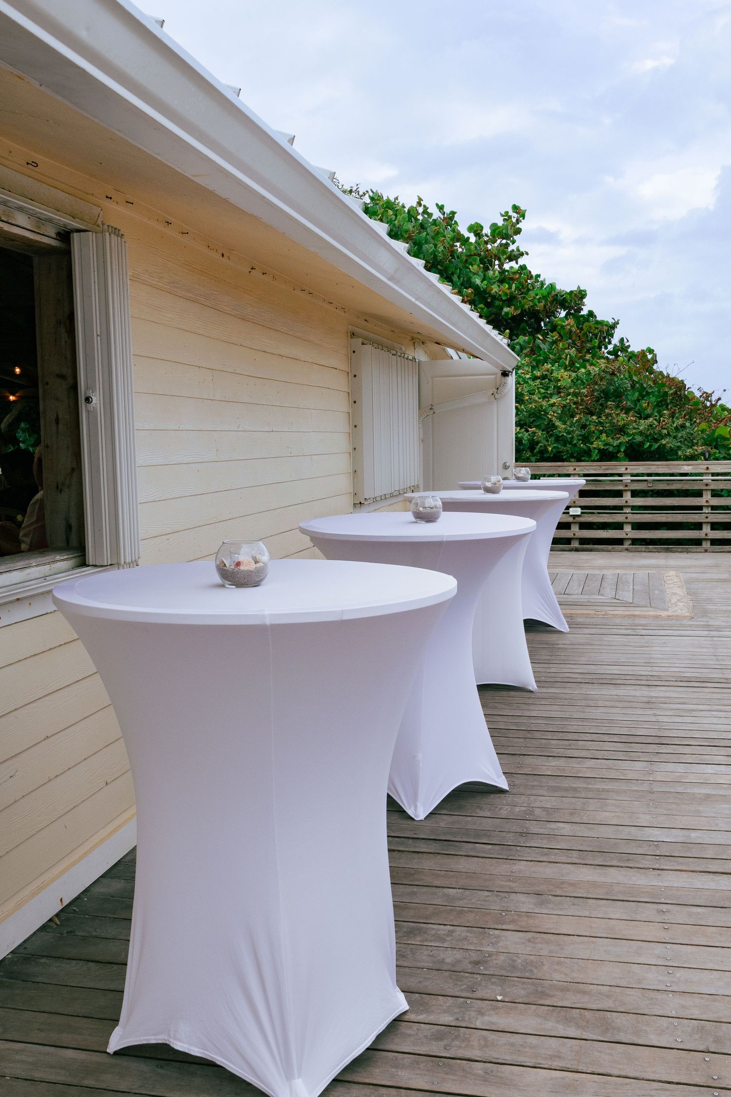 Row of white spandex cocktail tables on outdoor wooden deck at wedding venue.