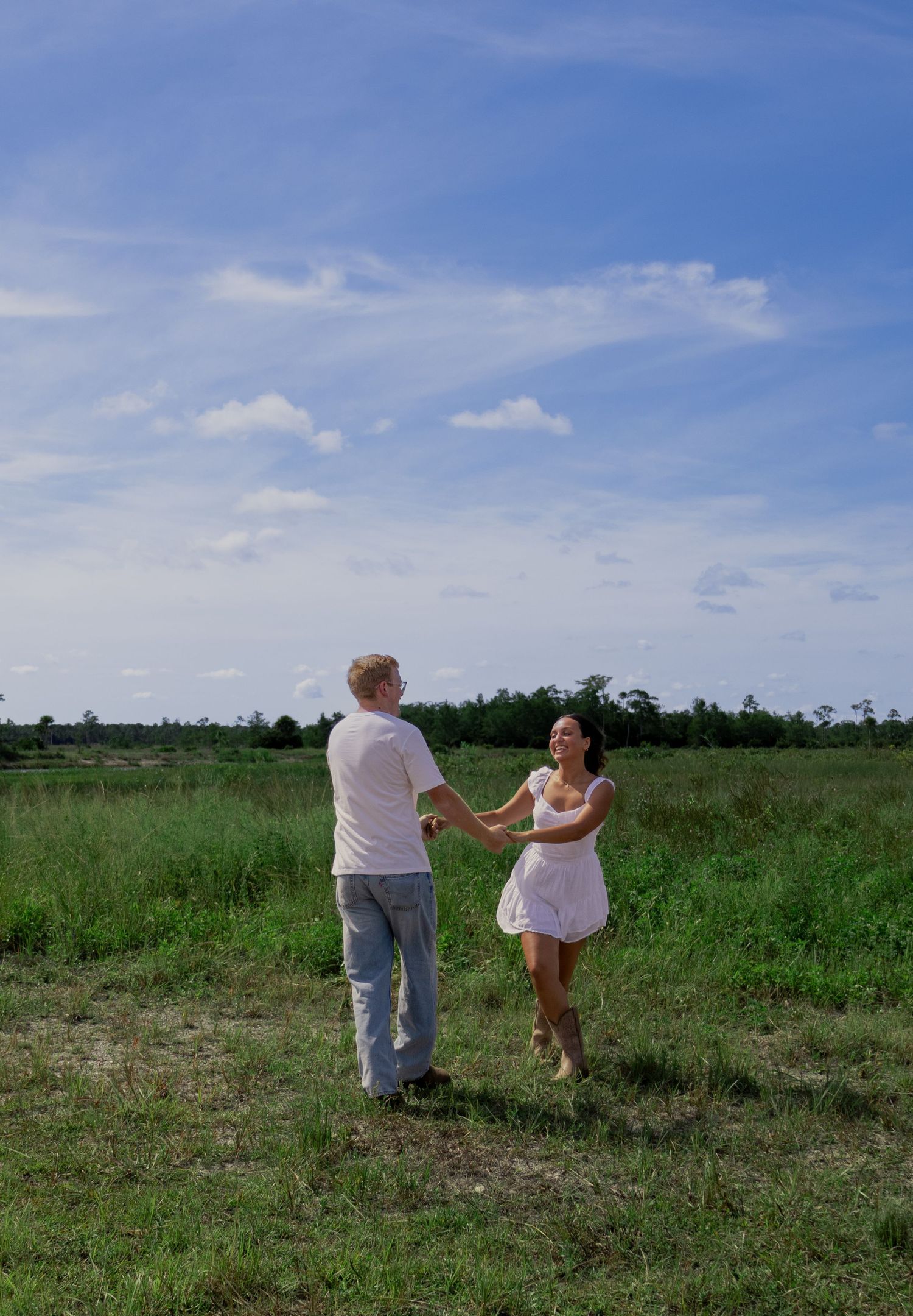 Happy couple holding hands and dancing together in an open green field under a blue sky.