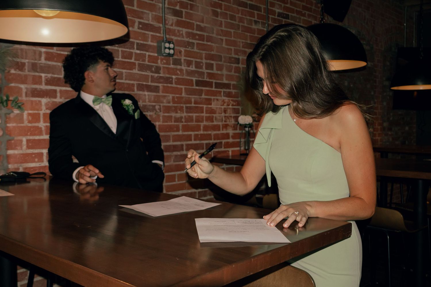 Wedding witness in sage dress signing marriage certificate with groom watching.