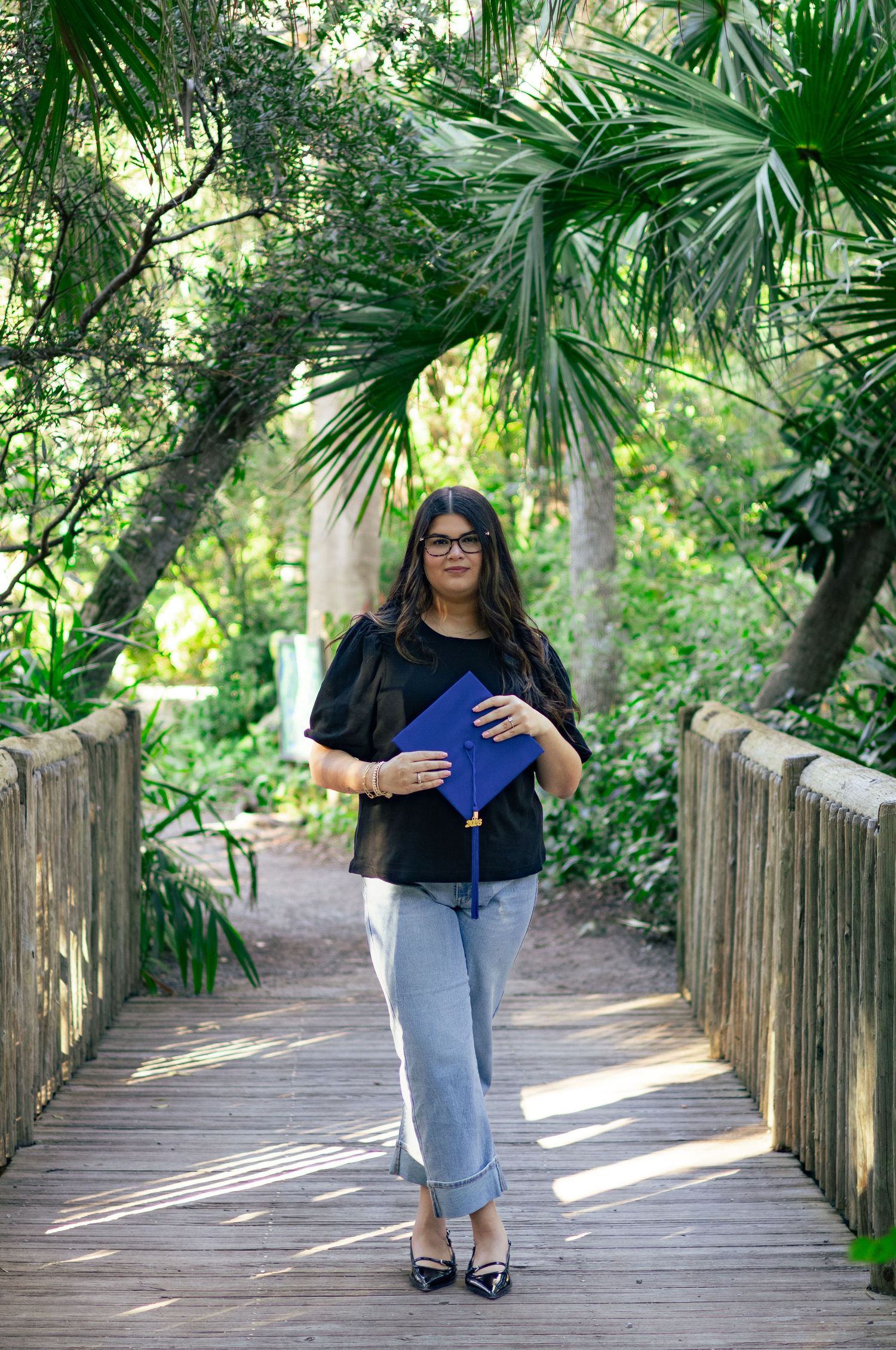 Graduate in black top holding blue diploma cover on wooden bridge path in tropical garden.