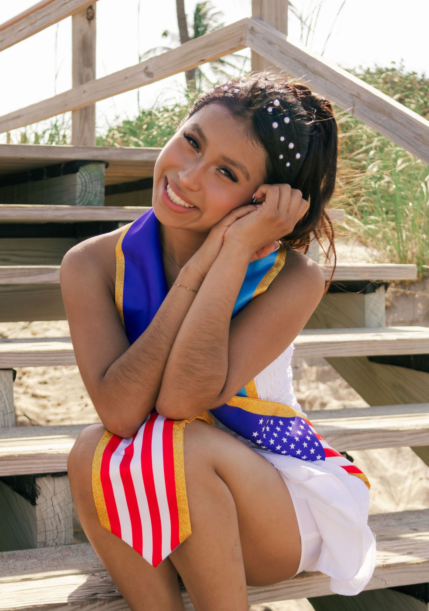 Smiling young Latina graduate sitting on beach boardwalk stairs with American flag graduation sash.