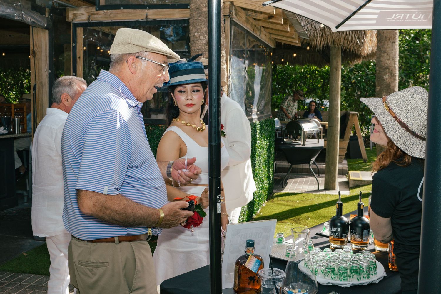 Guests in Derby attire conversing near a bar setup at an elegant outdoor Kentucky Derby event.