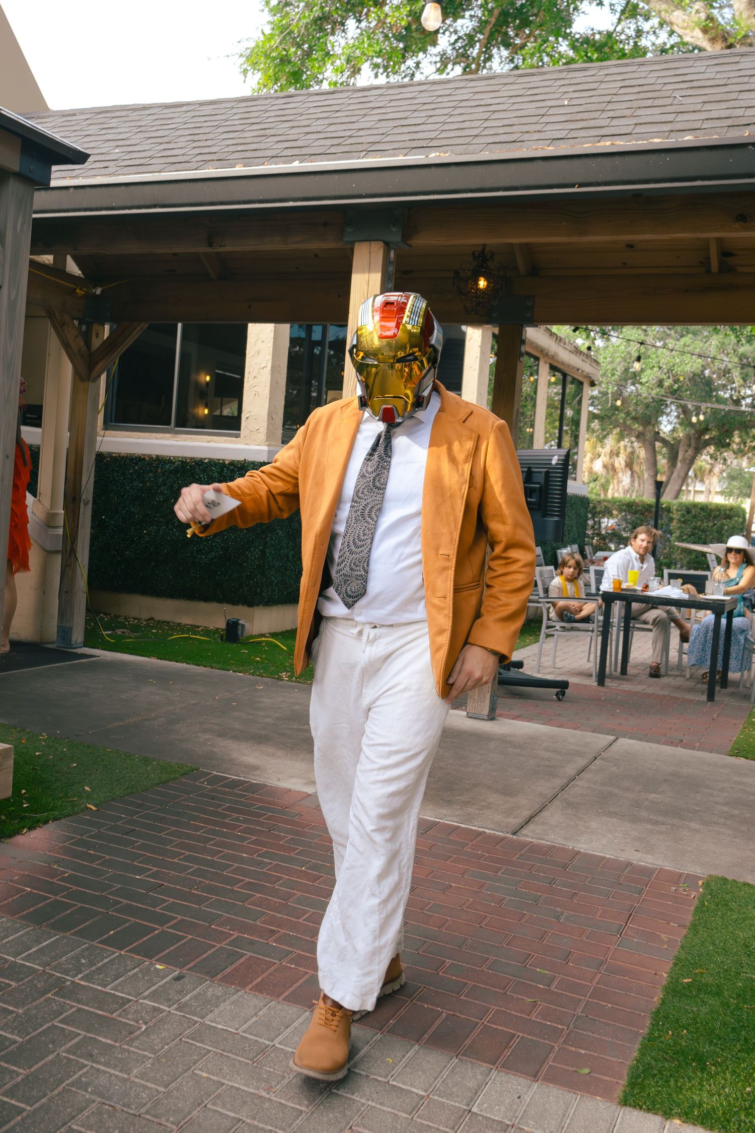 Man wearing gold wrestling mask, orange jacket, and white pants at an outdoor party event.