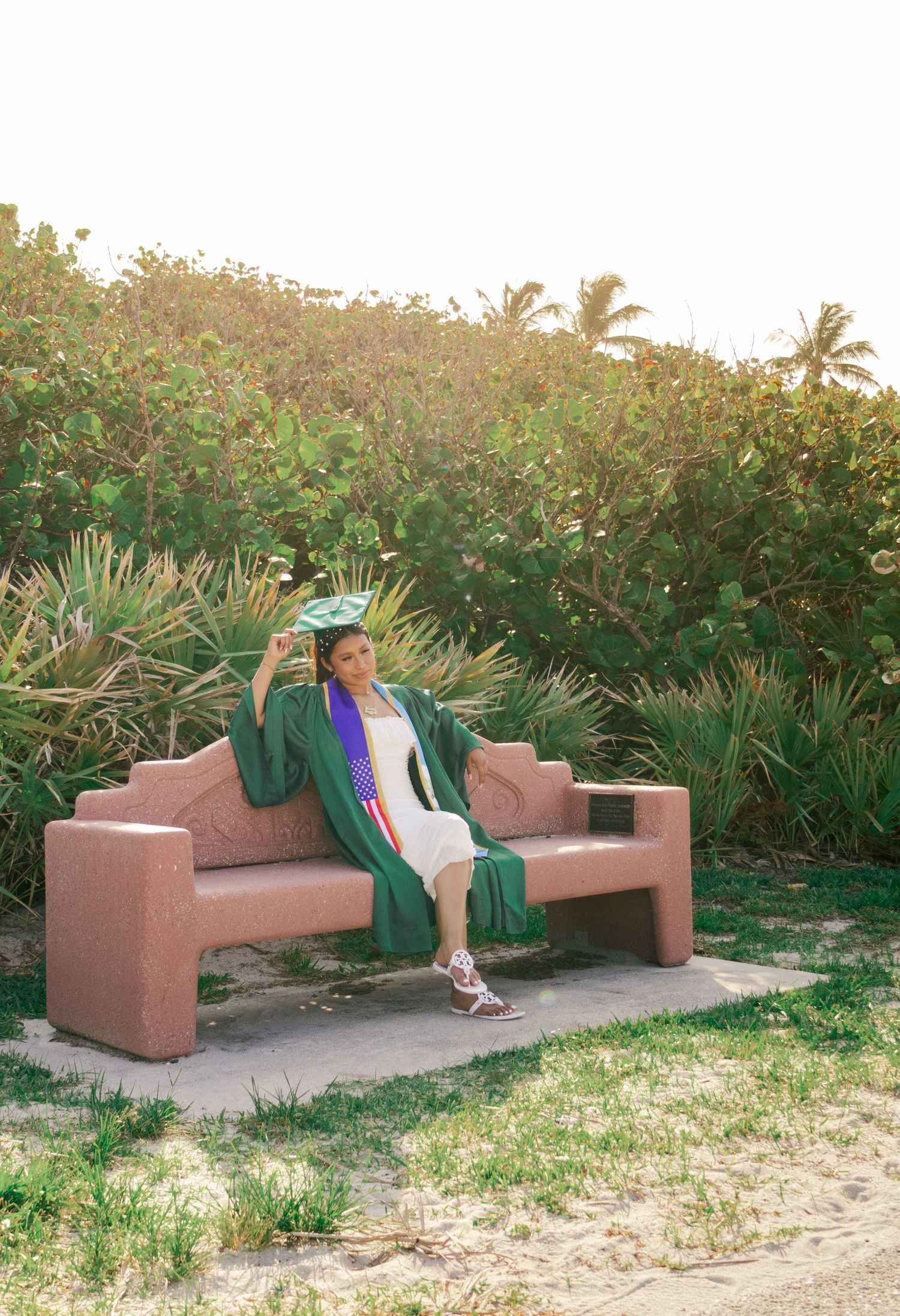 Female graduate in green cap and gown relaxes on a pink concrete bench surrounded by tropical greenery.