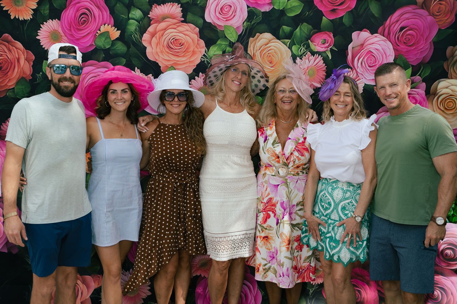 Group of seven friends posing in front of floral backdrop, women wearing colorful Kentucky Derby-style hats.