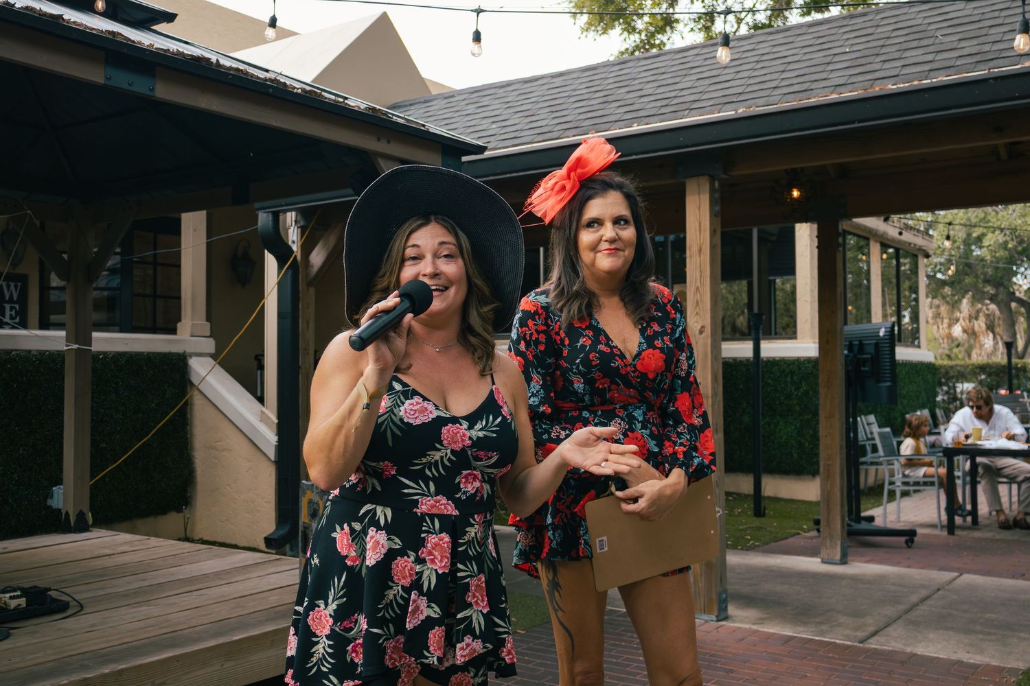 Two women in floral dresses host outdoor event, one speaking into microphone wearing black hat, other in red fascinator.