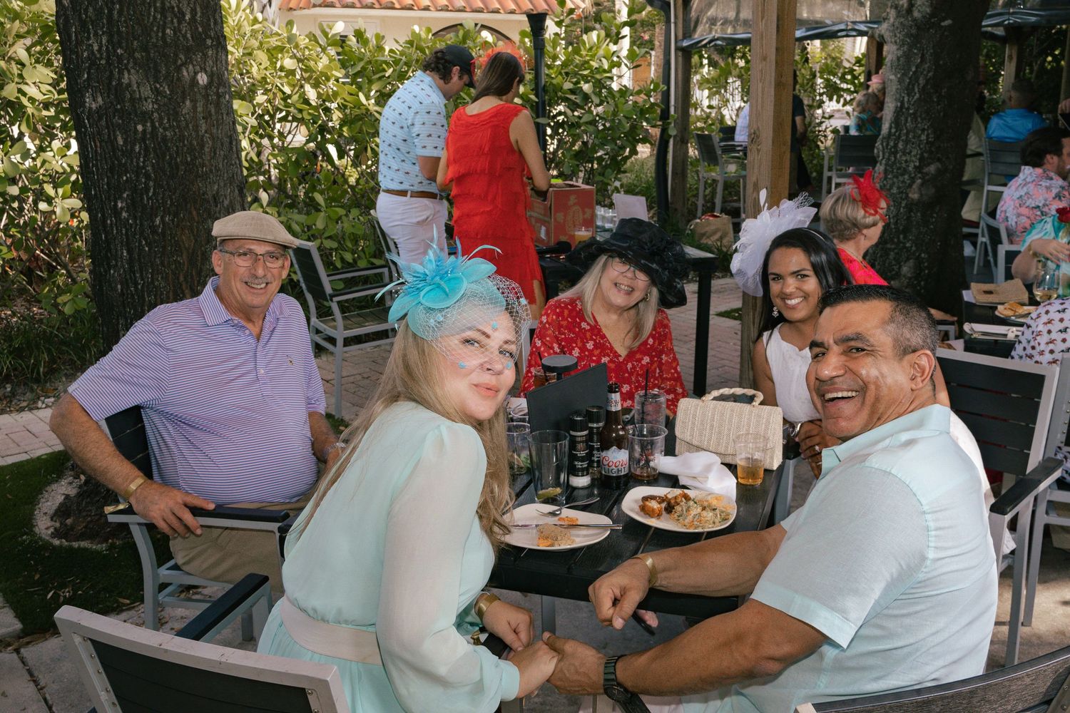 Group of friends wearing decorative hats enjoy food and drinks at an outdoor Kentucky Derby watch party.