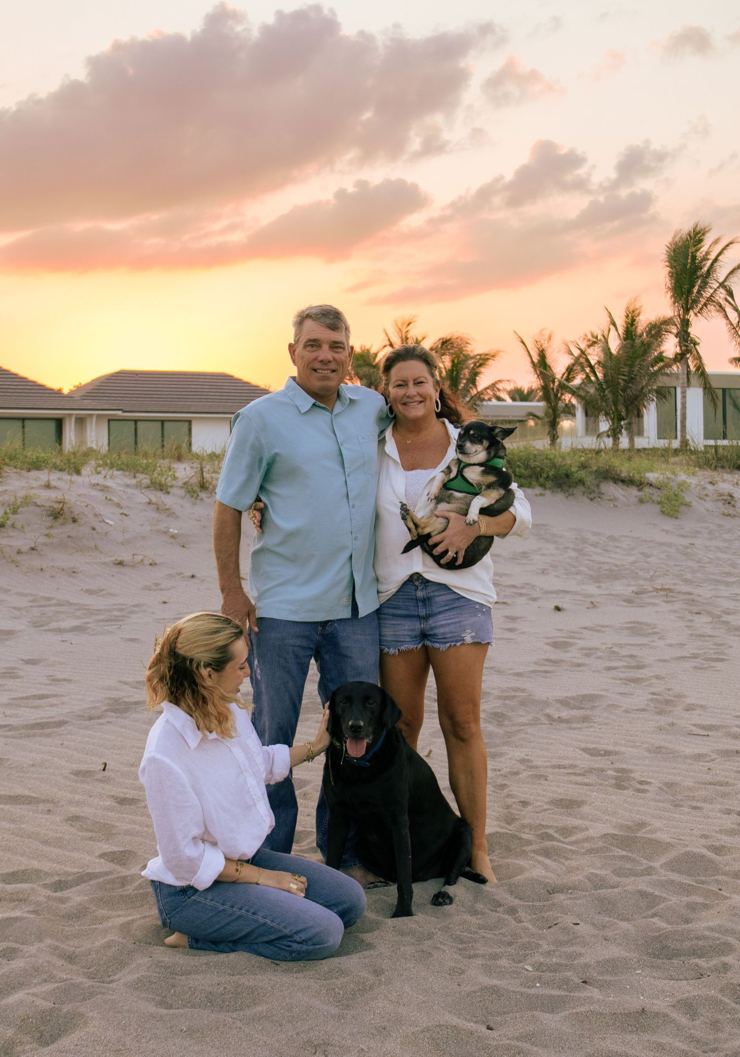 Family of three poses with two dogs on sandy beach at golden sunset with palm trees and beach houses in background.