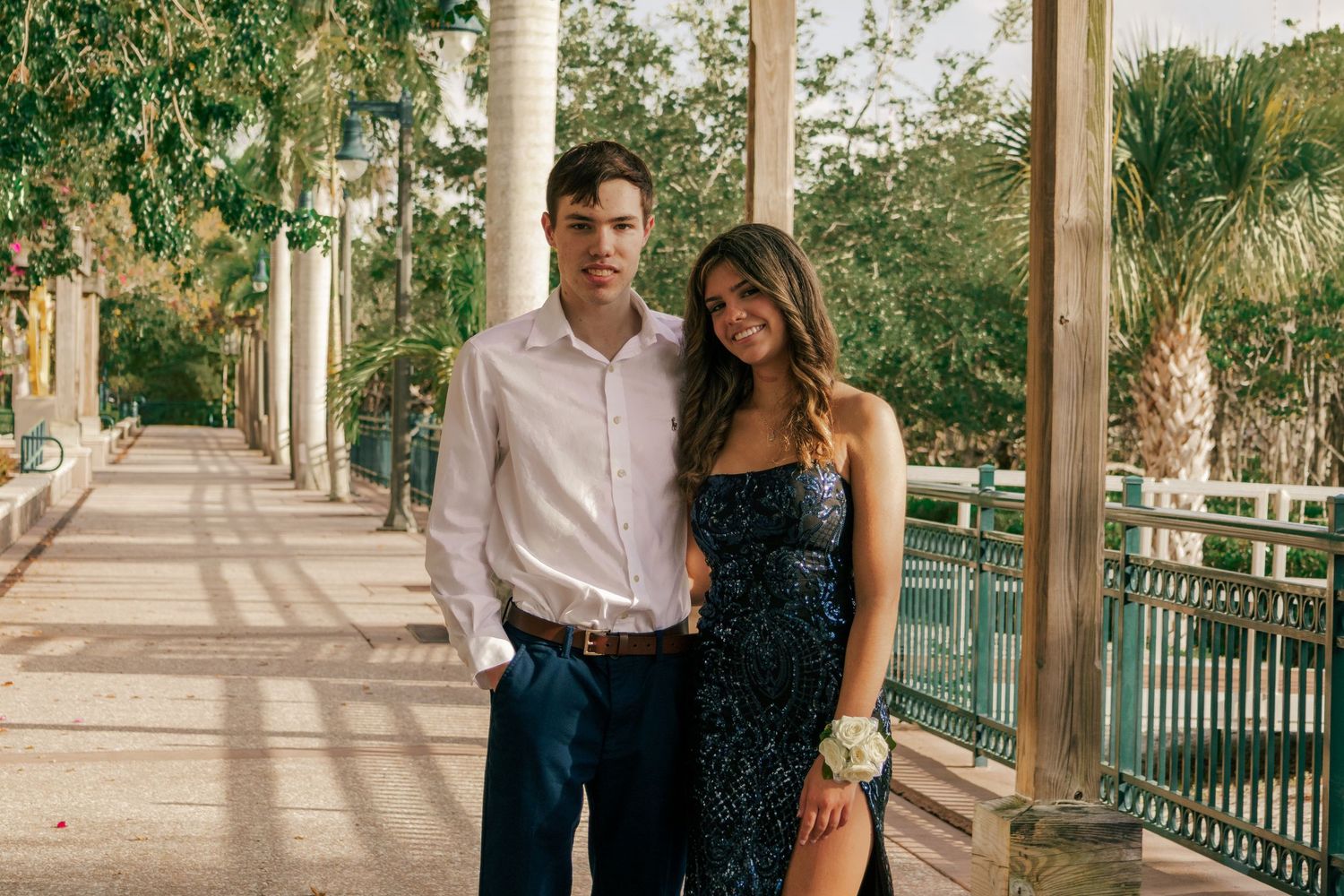 A young couple dressed in formal prom attire posing on an elegant columned walkway surrounded by lush greenery.