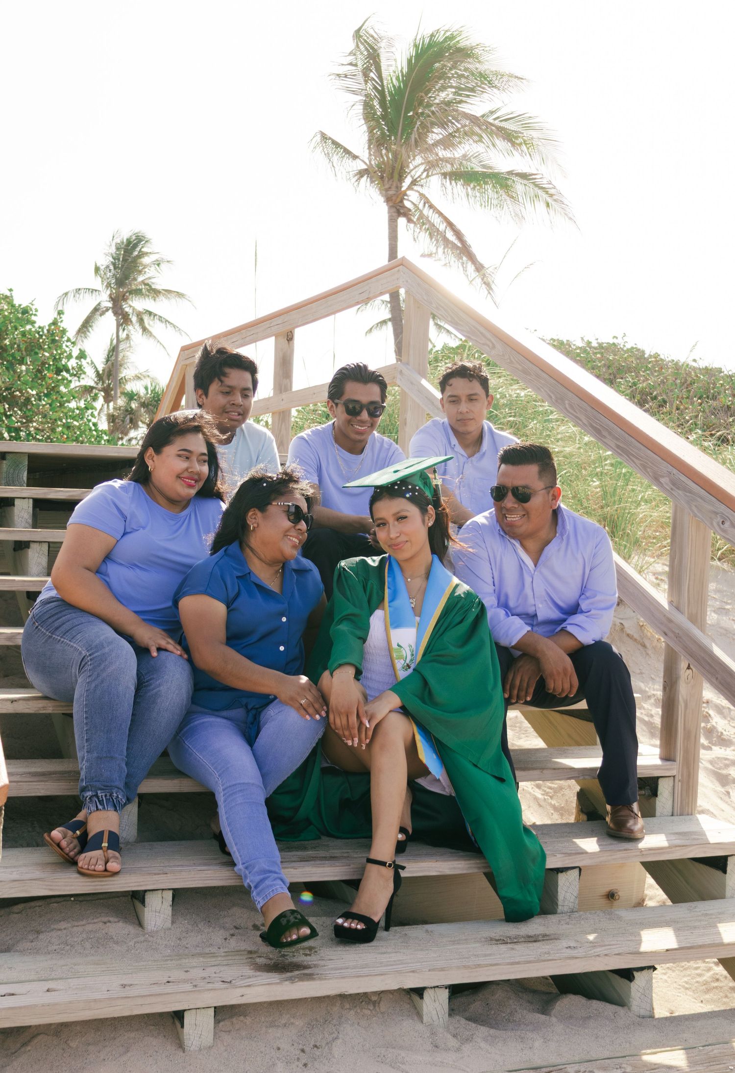 Graduate in green cap and gown poses with family on beach boardwalk stairs near palm trees.
