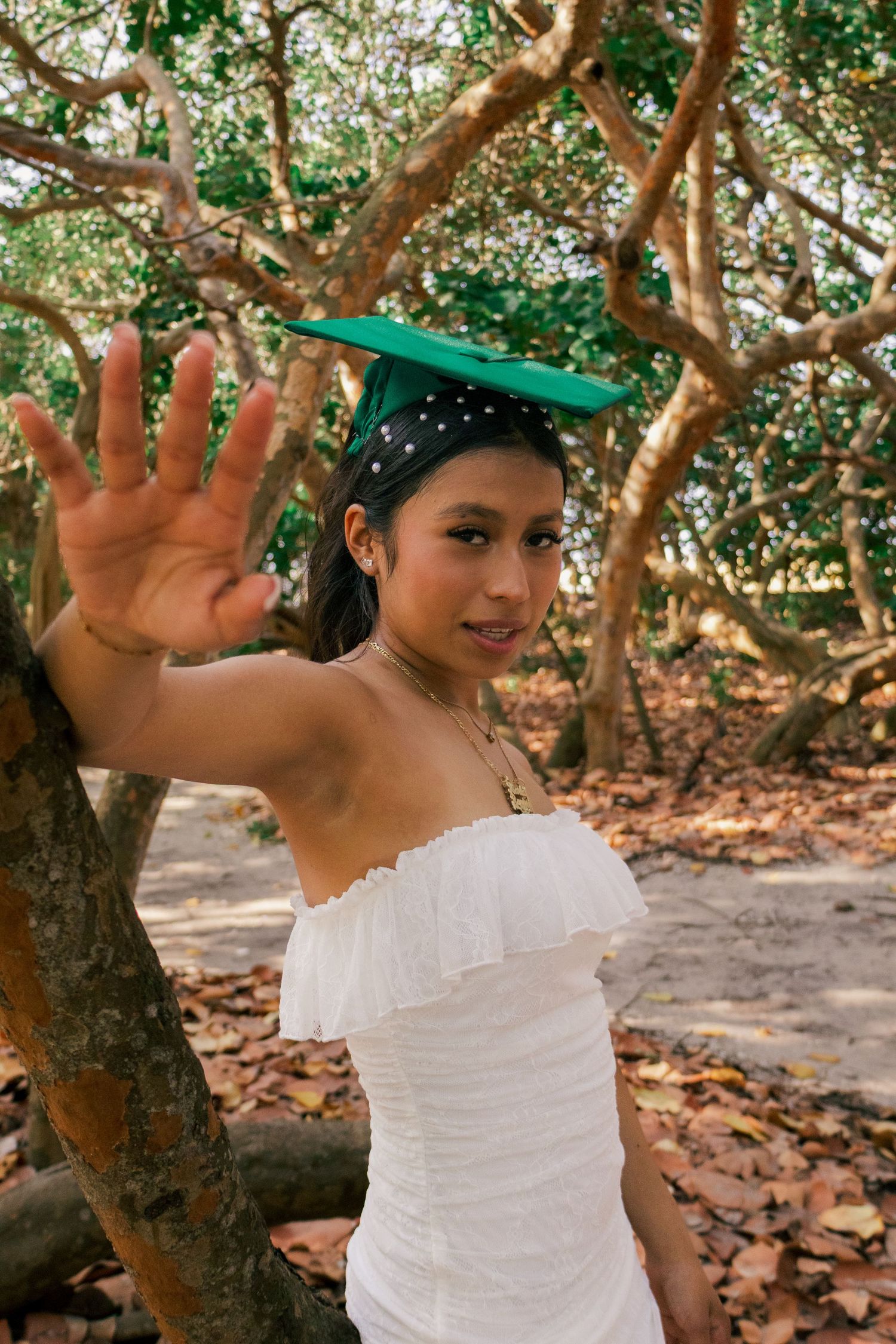 Graduate in white off-shoulder dress and green cap playfully reaching toward camera in a scenic wooded area.