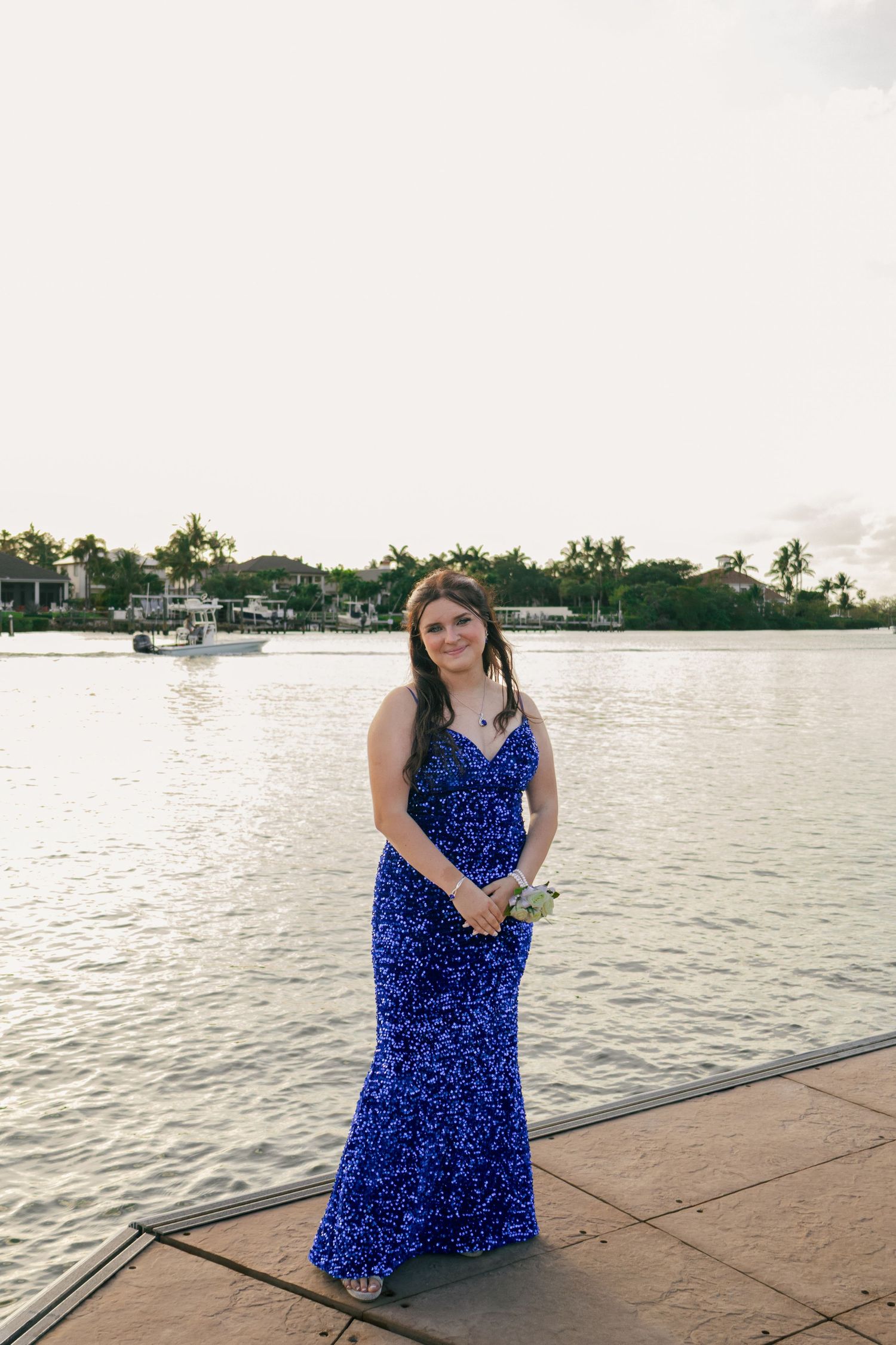A teenage girl in a royal blue sequin prom dress poses holding a corsage on a Florida waterfront dock.
