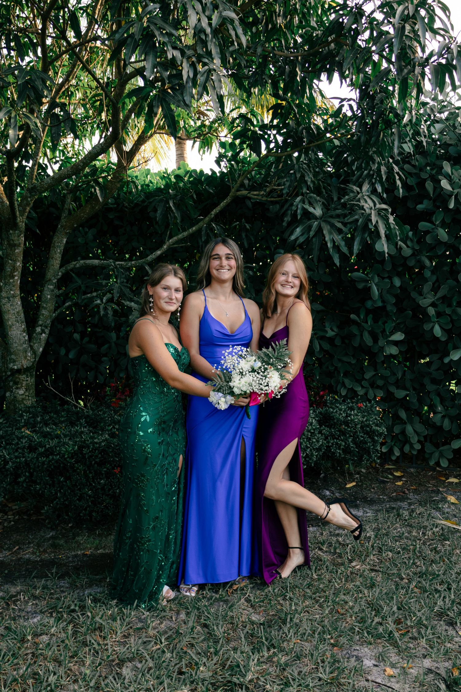 Three prom girls pose outdoors in green, blue, and purple gowns holding floral bouquets under trees.