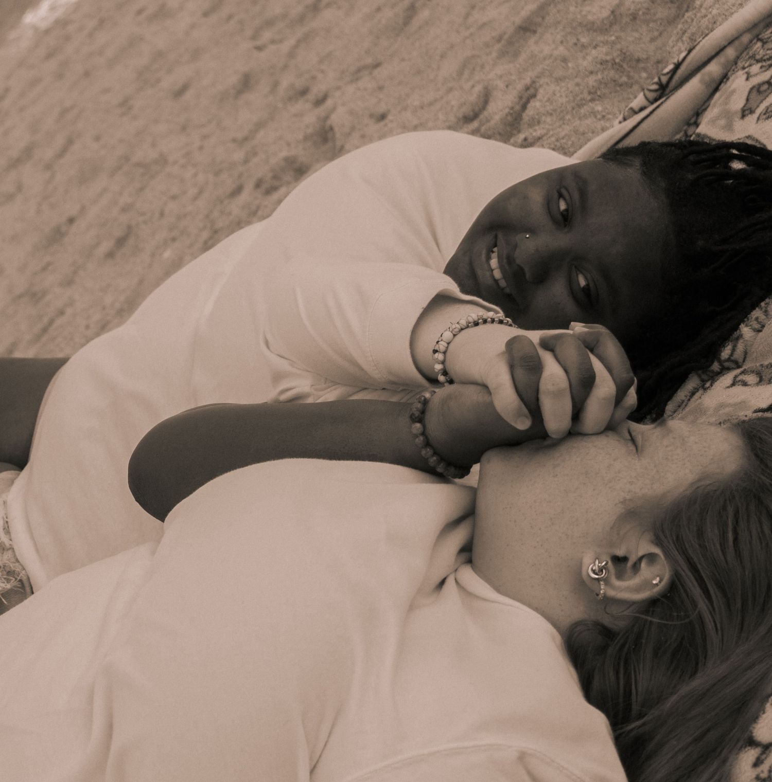 Two women lying on sand, smiling at each other in sepia tone.