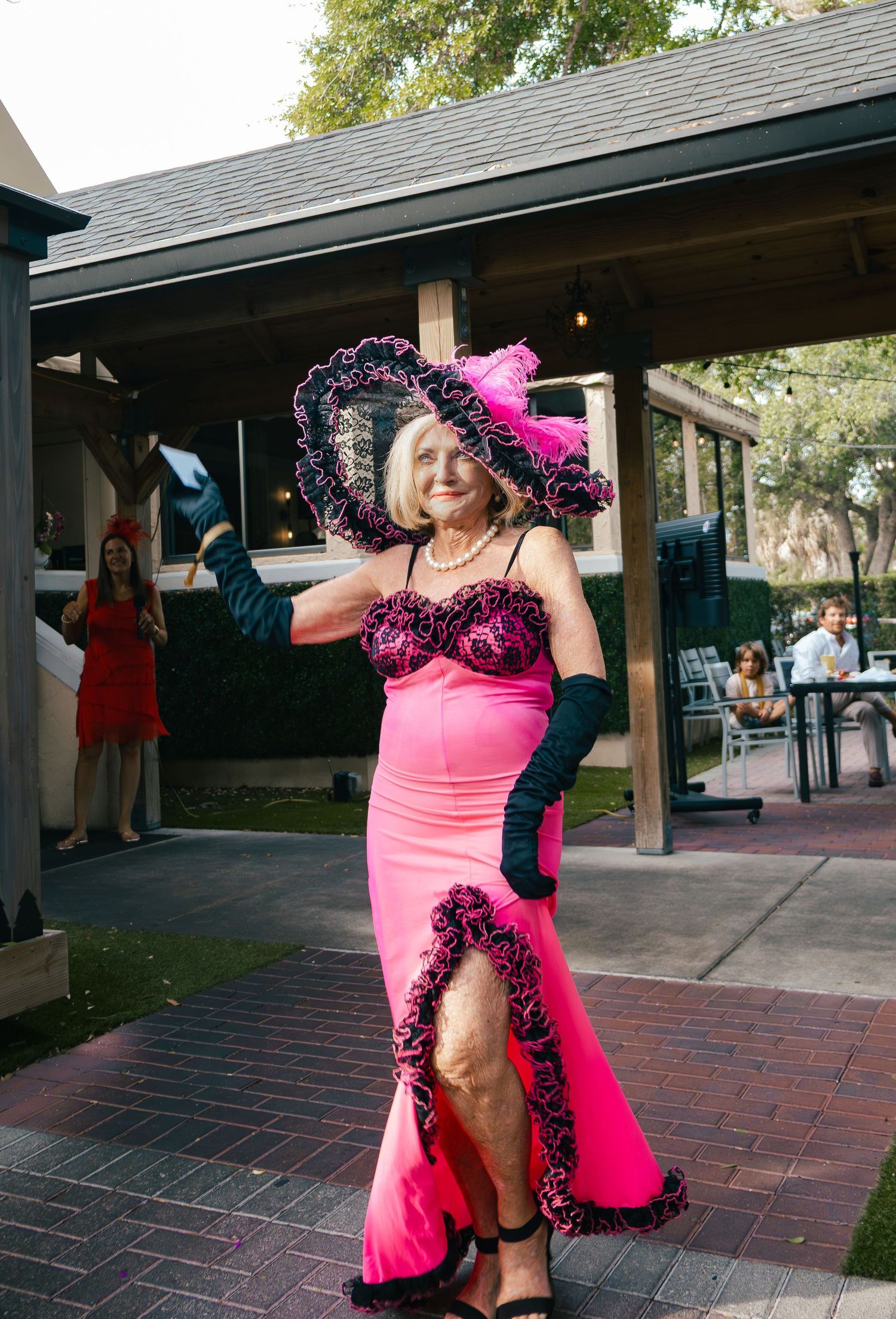 Elderly woman in hot pink gown with black lace trim, long gloves, and matching wide-brim hat posing at outdoor event.